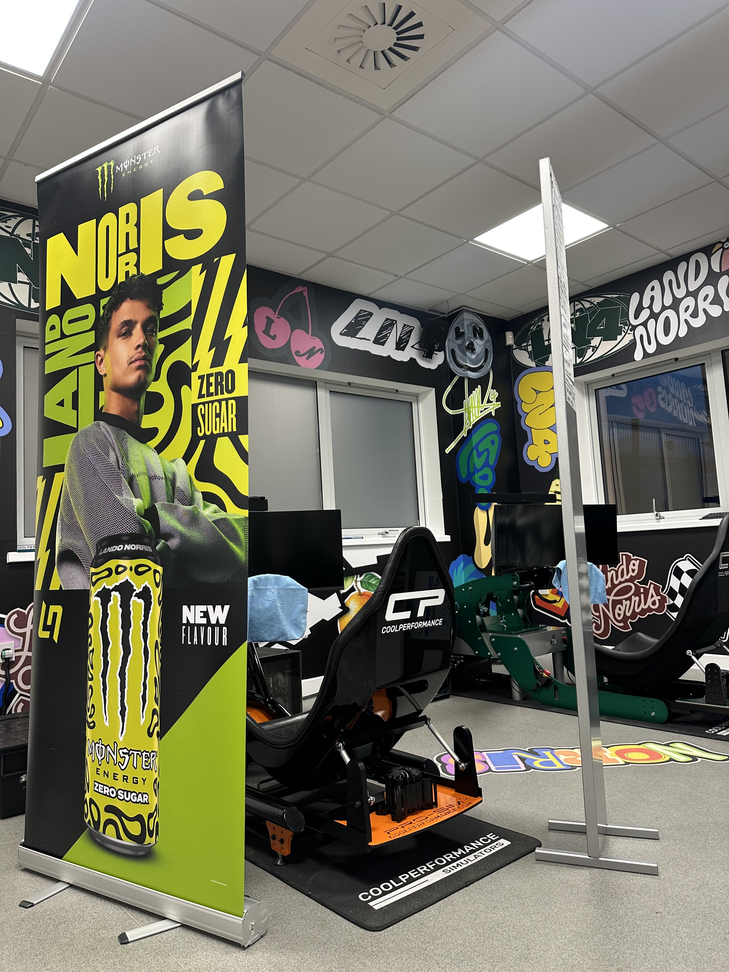 Indoor display of a racing simulator with branding for Monster Energy and Lando Norriss. The setup includes a racing seat, steering wheel, and pedals, with colorful logos and posters on the walls.