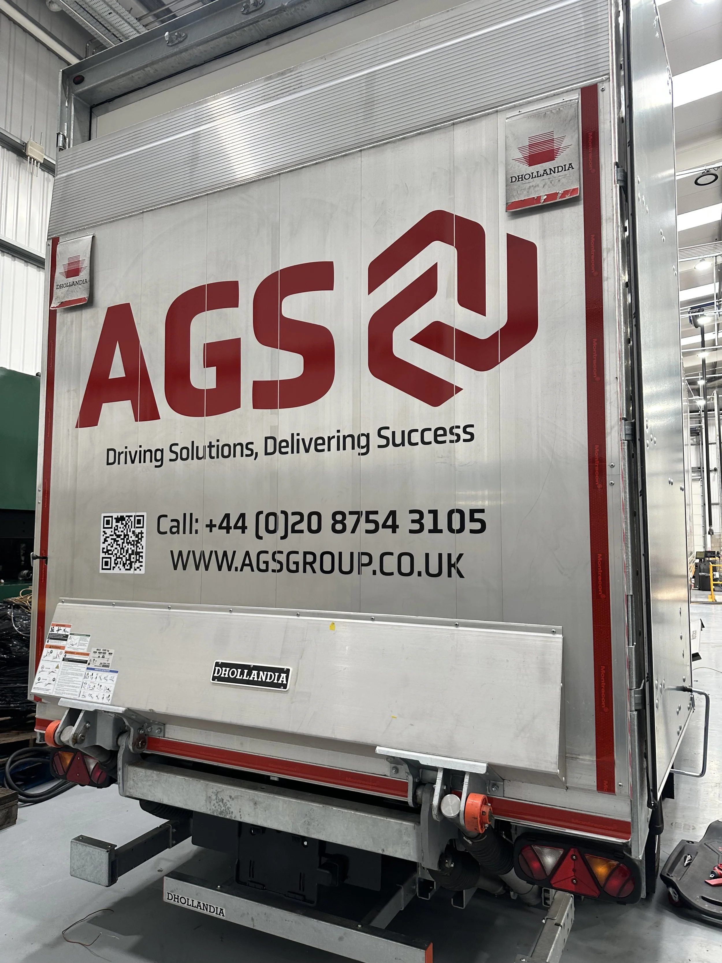 Large metallic vehicle with branding and contact information for AGS Group, including the slogan "Driving Solutions, Delivering Success," in industrial setting.
