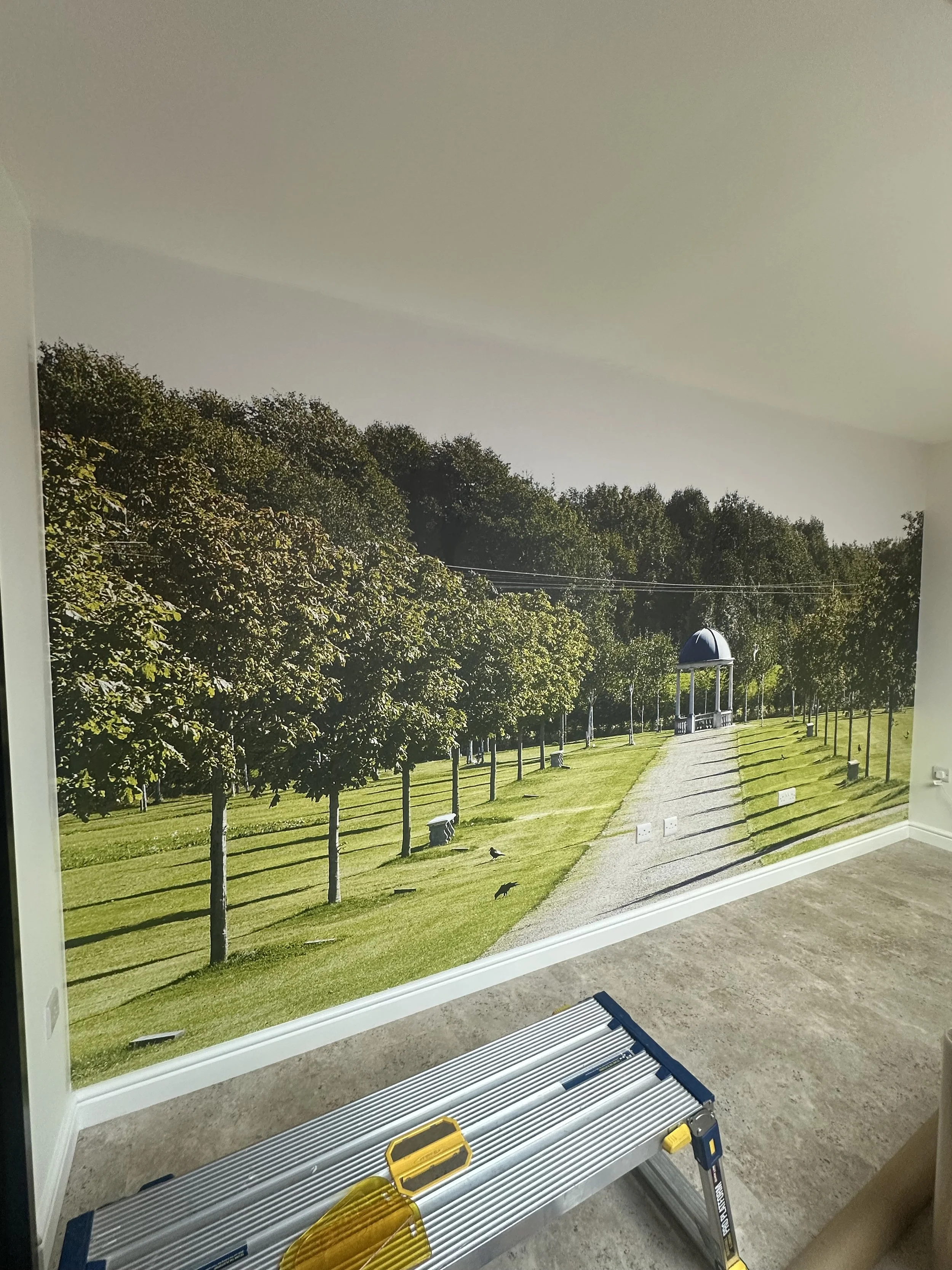 Interior wall with a large one piece wallpaper of a mural of a park scene featuring trees, a gazebo, a grassy area, and a path, with a ladder and toolbox in the foreground.