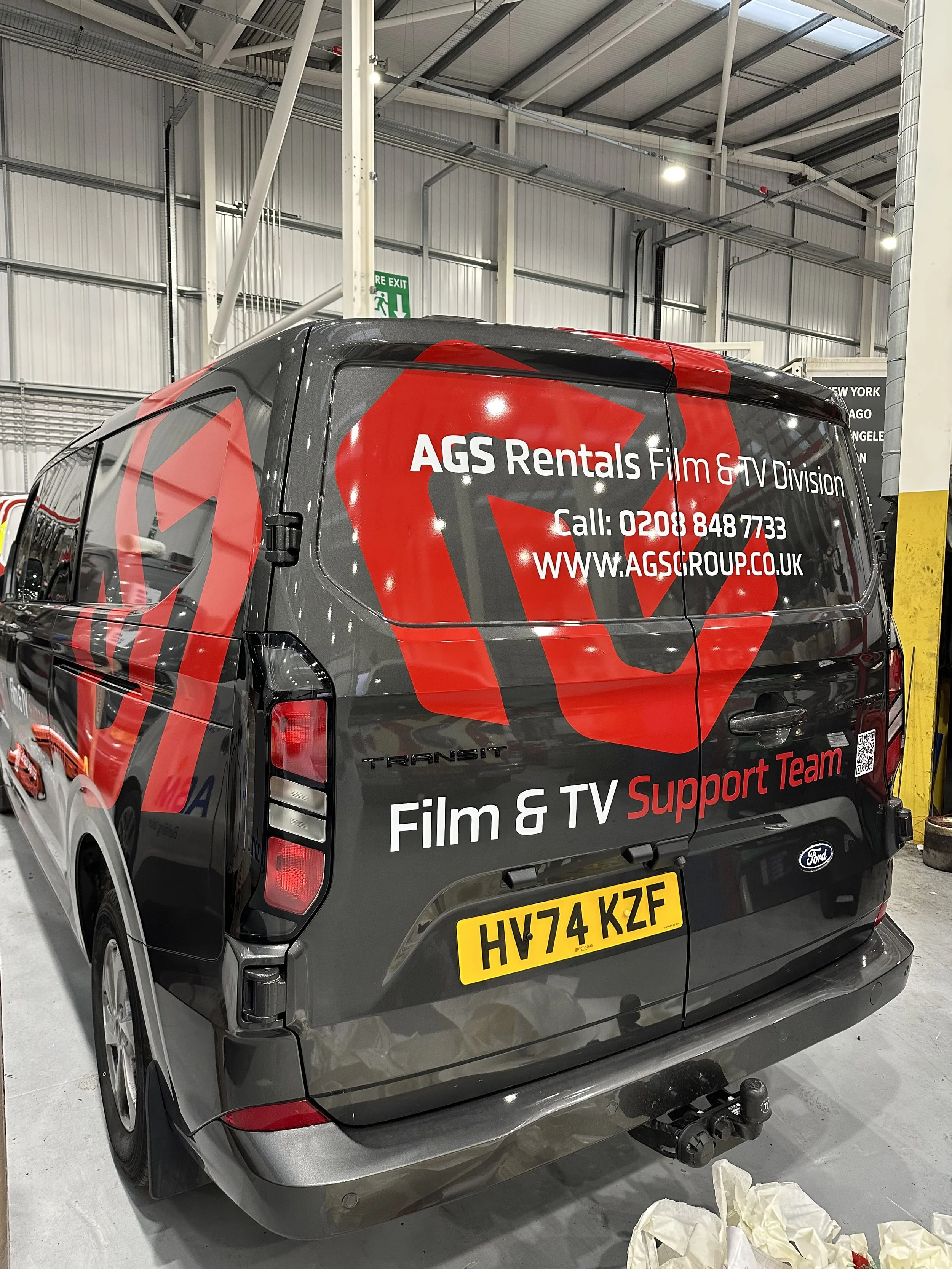 A dark gray Ford Transit van in an indoor warehouse or garage. The van has large red graphic text advertising AGS Rentals Film & TV Division, including contact information and website. The rear of the van shows a UK license plate and a trailer hitch.