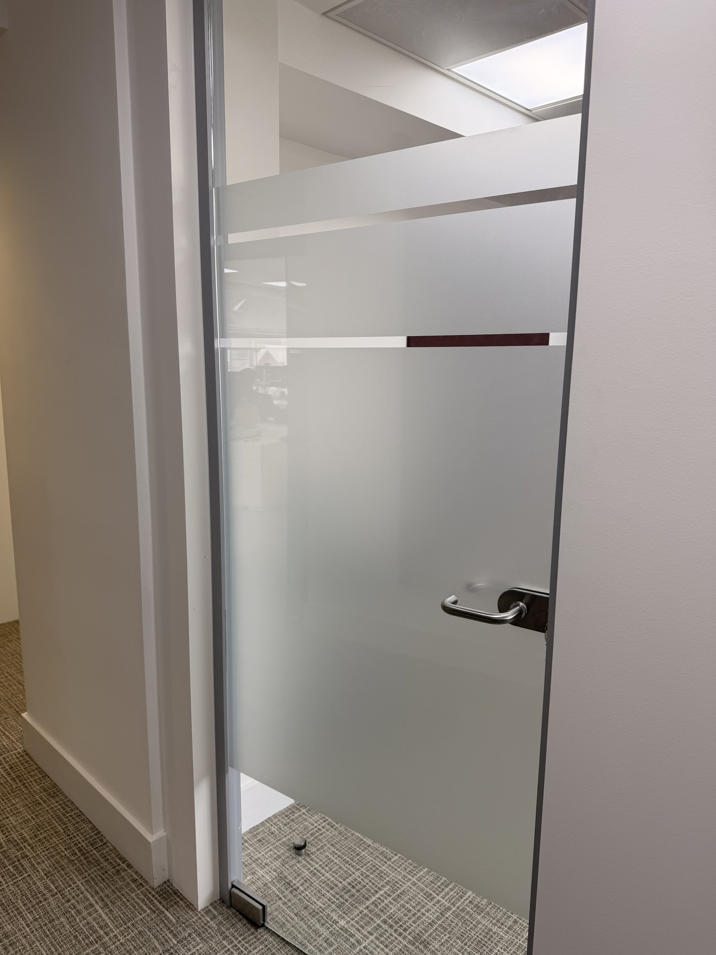 An open glass door with a metal handle leading to a small, enclosed room with frosted glass walls and a ceiling vent.