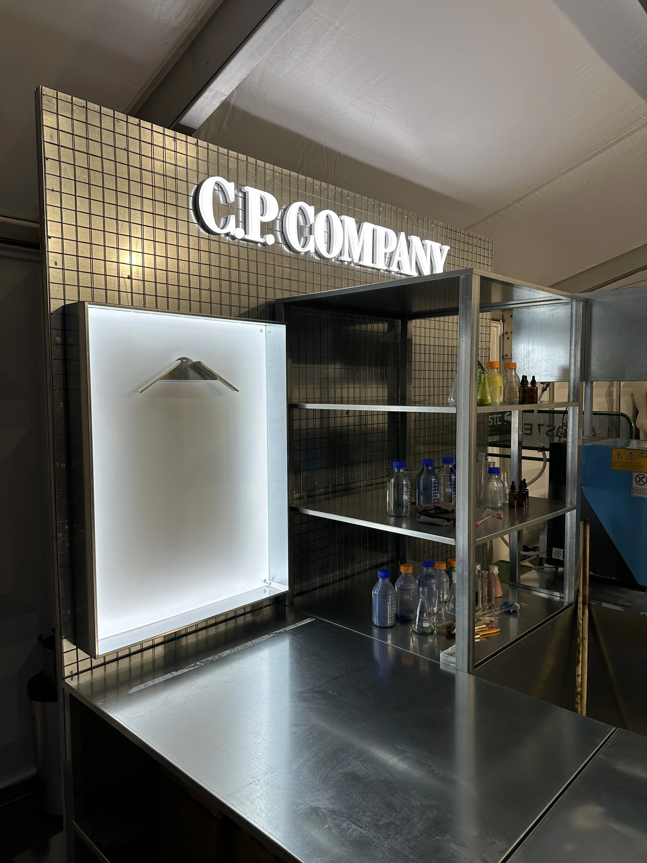 A stainless steel event setup, countertop with shelves holding glass bottles and laboratory equipment, with a grid-patterned wall and illuminated sign that reads 'C.P. COMPANY' in the background.