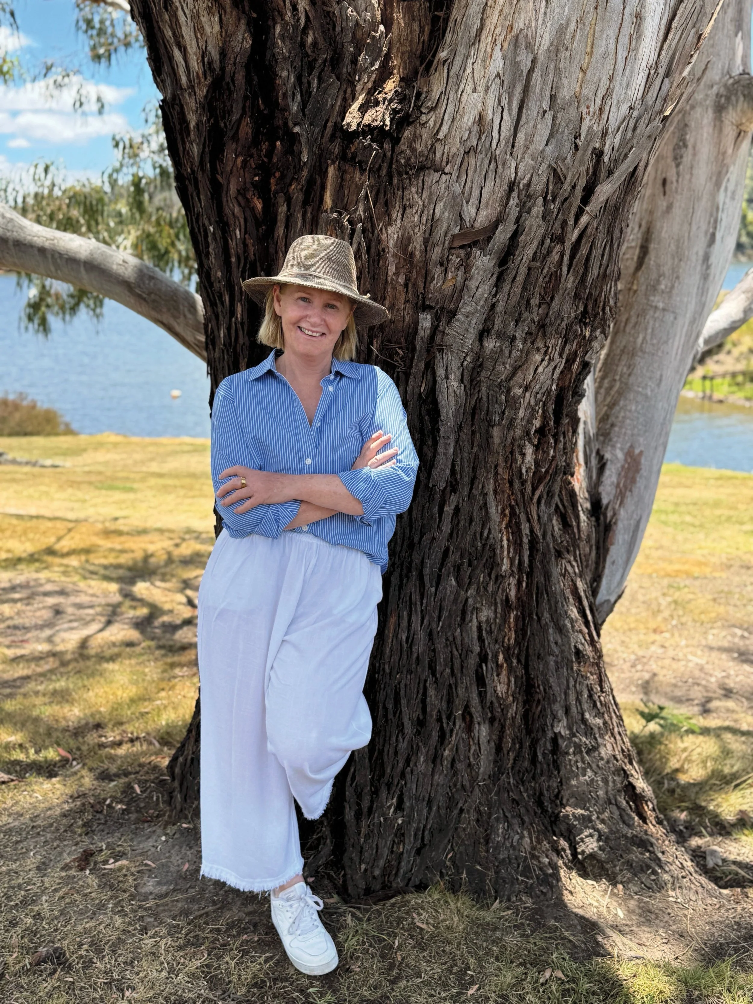 A woman standing next to a large tree near a body of water, wearing a blue striped shirt, white wide-leg pants, white sneakers, and a wide-brimmed hat, smiling at the camera.