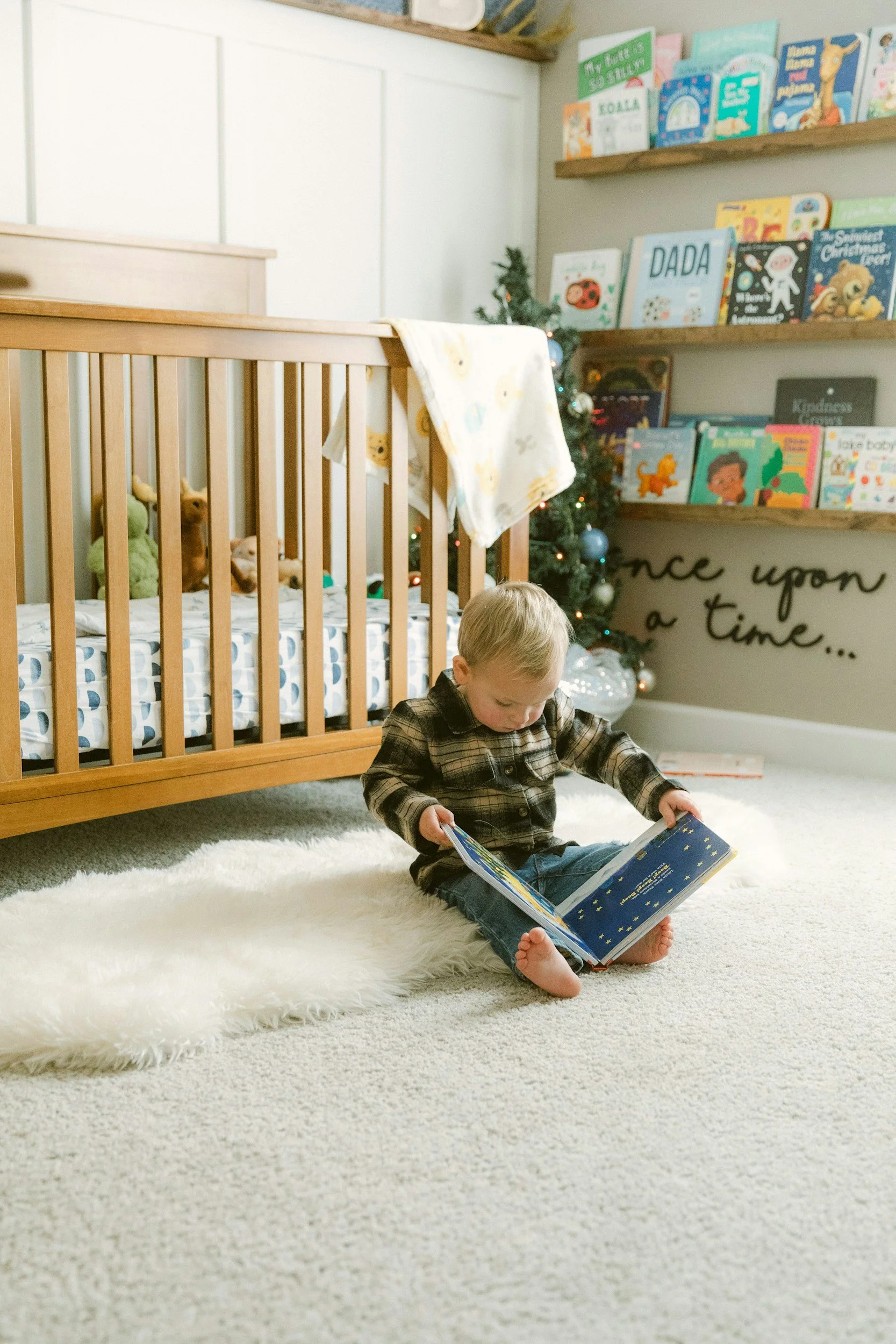 Small boy in a flannel top and jean sitting on  a sheepskin rug next to his cot reading a picture book