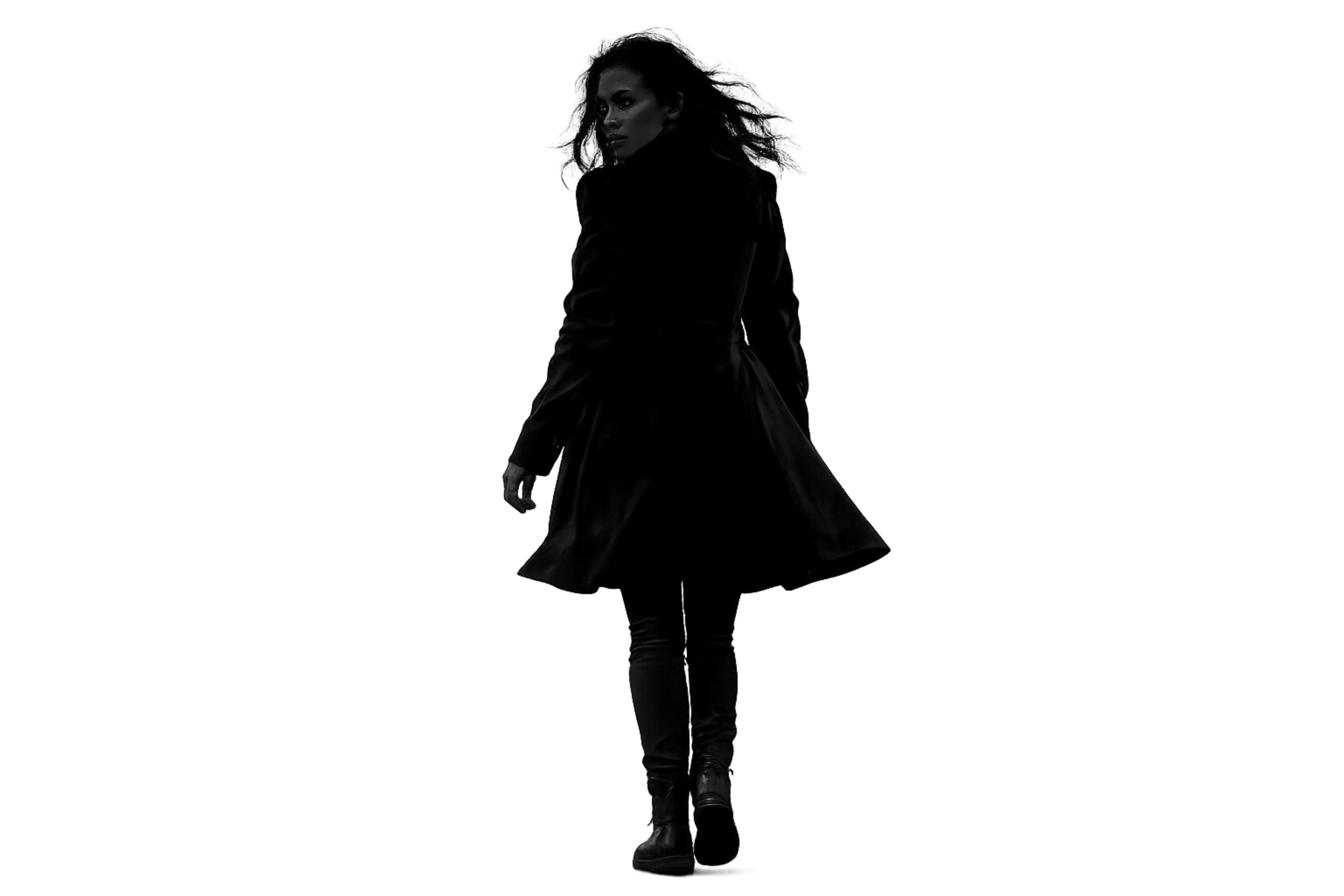 A woman with curly hair wearing a long black coat and black boots, walking away in a dark, neon-lit cityscape.