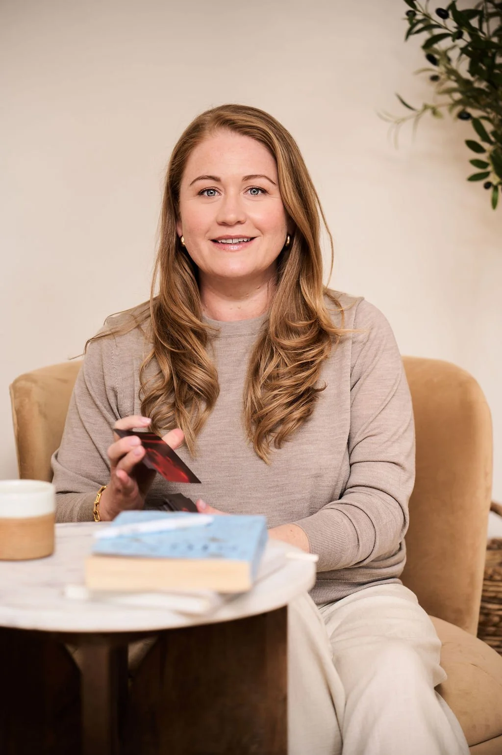 A woman with long, wavy, light brown hair, wearing a beige top, sitting on a beige upholstered chair, smiling, holding playing cards, with a table in front of her that has a blue book, a white mug, and a small bowl. There is a plant with green leaves in the background.