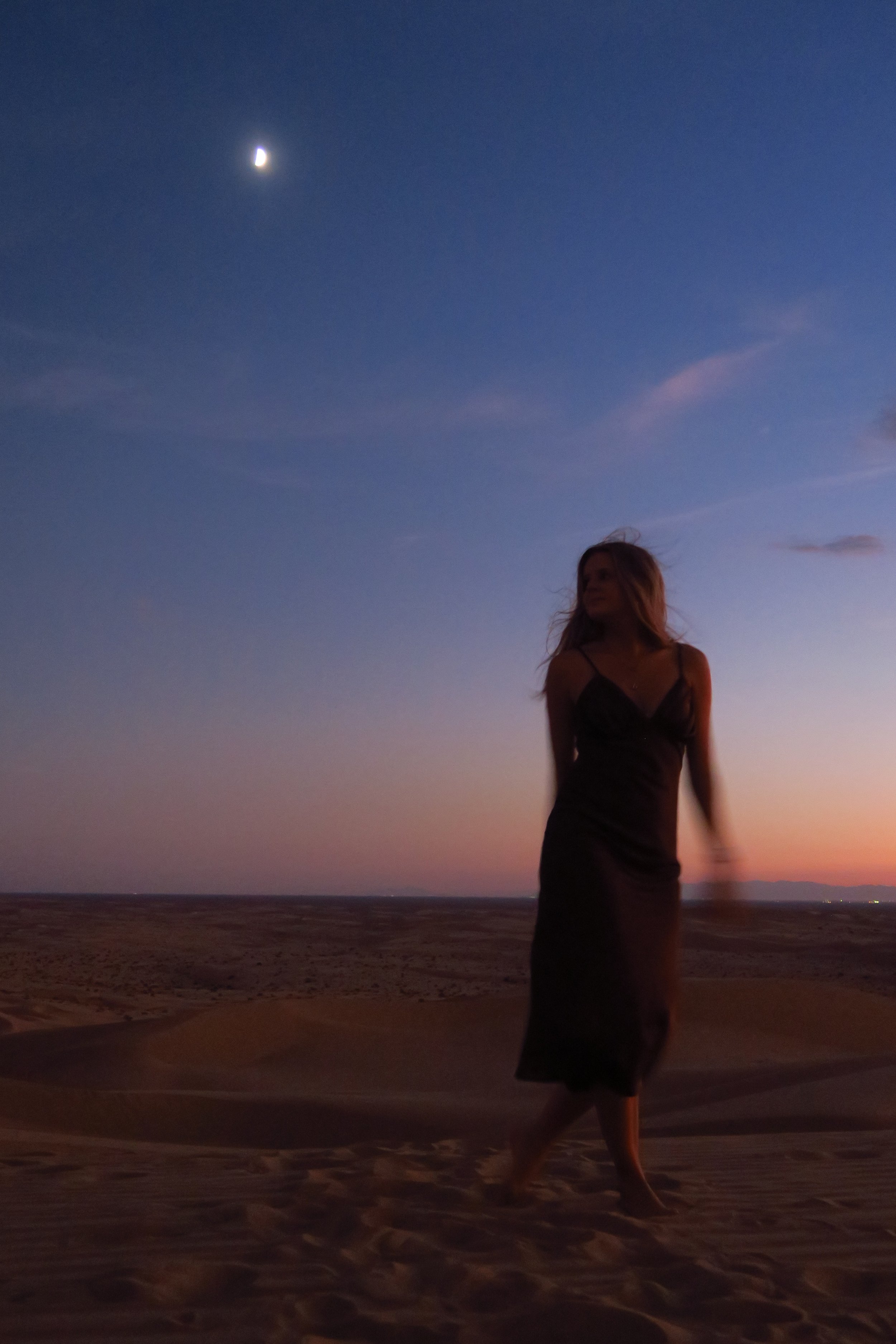 A woman in a black dress walking on sand dunes at dusk with a crescent moon in the sky.
