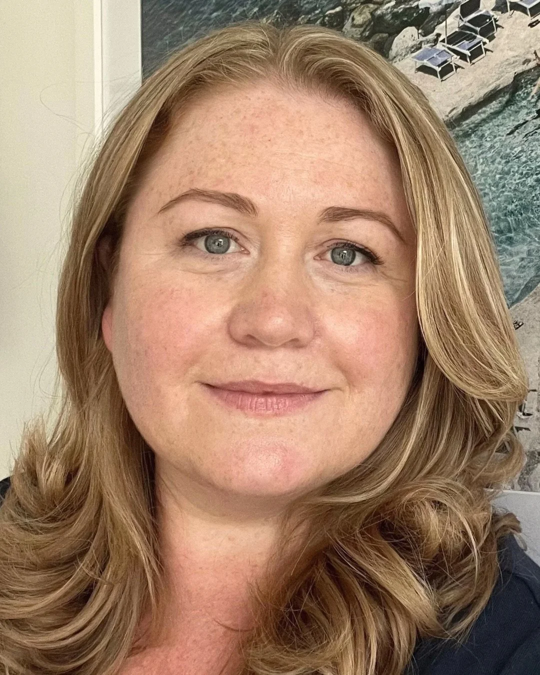 Close-up of a woman with shoulder-length wavy blonde hair, blue eyes, and fair skin, smiling softly at the camera with a beach scene poster in the background.