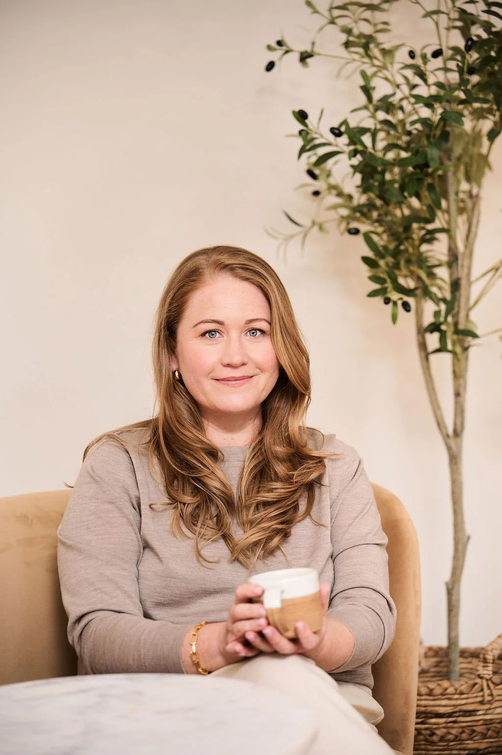 A young woman with long, wavy red hair, wearing a beige sweater, sitting on a beige couch, holding a mug wrapped in a wooden sleeve, smiling at the camera, with a tall potted plant behind her.