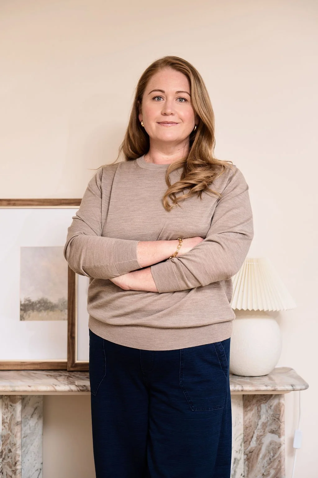A woman with long, wavy, light brown hair standing with her arms crossed in front of a beige wall. She is wearing a beige long-sleeve shirt and dark blue pants. Behind her is a marble table with a framed picture and a white table lamp with a pleated shade.