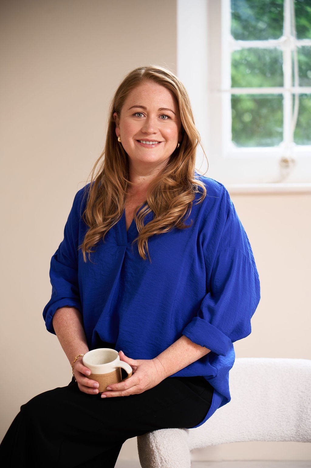 Woman with long wavy brown hair, wearing a blue blouse, sitting on a white chair, holding a coffee mug, smiling in front of a window with greenery outside.