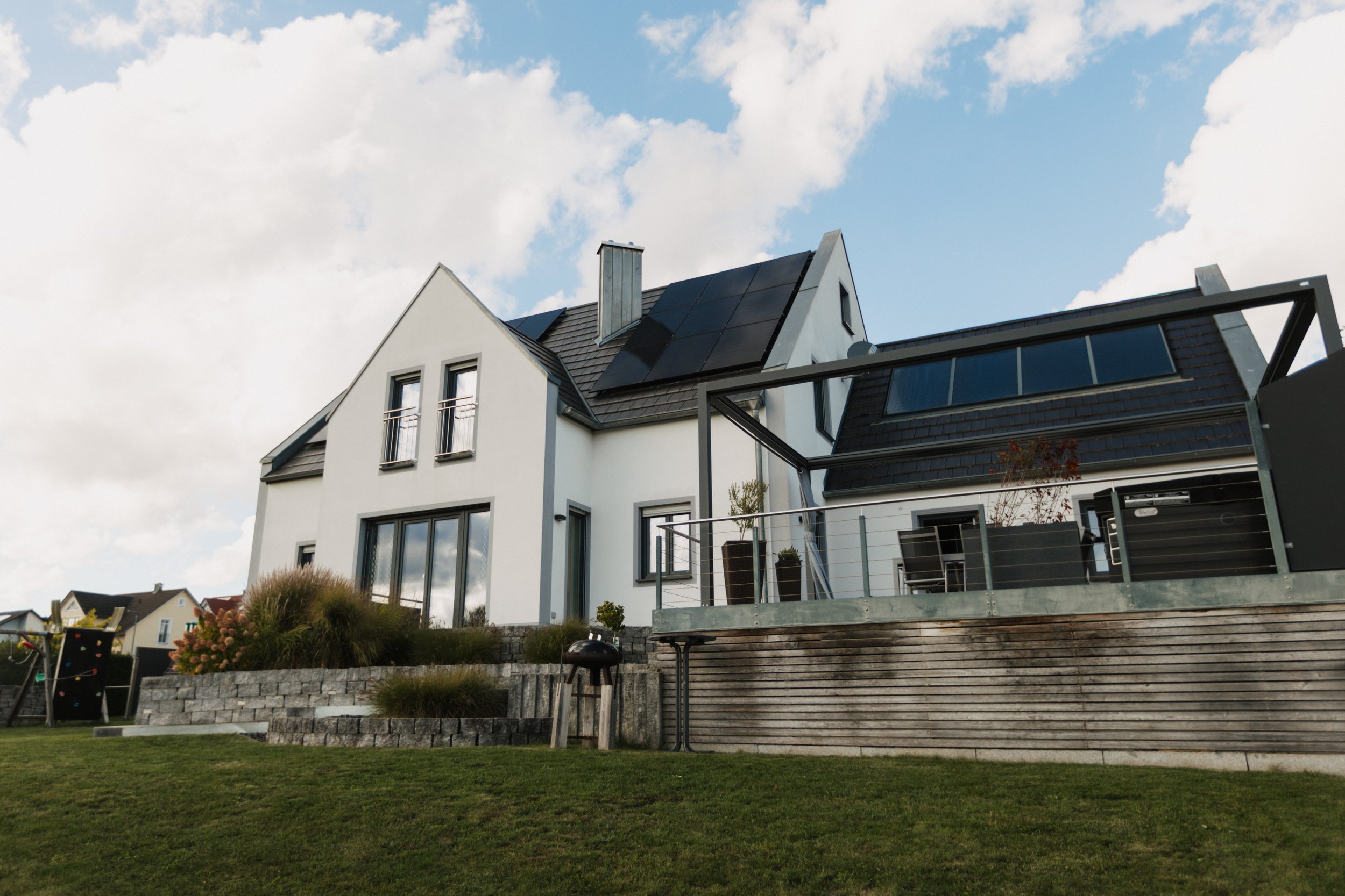 Modernes weißes Haus mit großen Fenstern und Solardach, Umgebung mit Garten, Rasen und Terrasse, unter blauem Himmel mit Wolken.