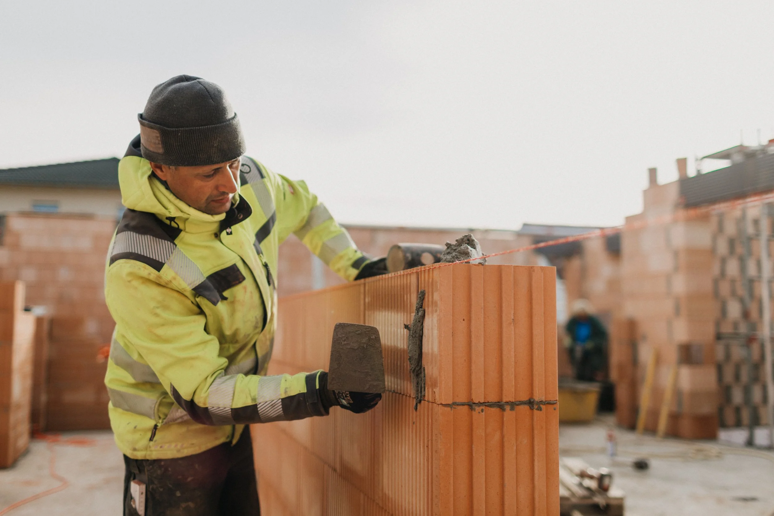 Bauer richtet Ziegelmauer auf einer Baustelle mit Bauhelfer im Hintergrund, der mit Schutzhelm arbeitet.