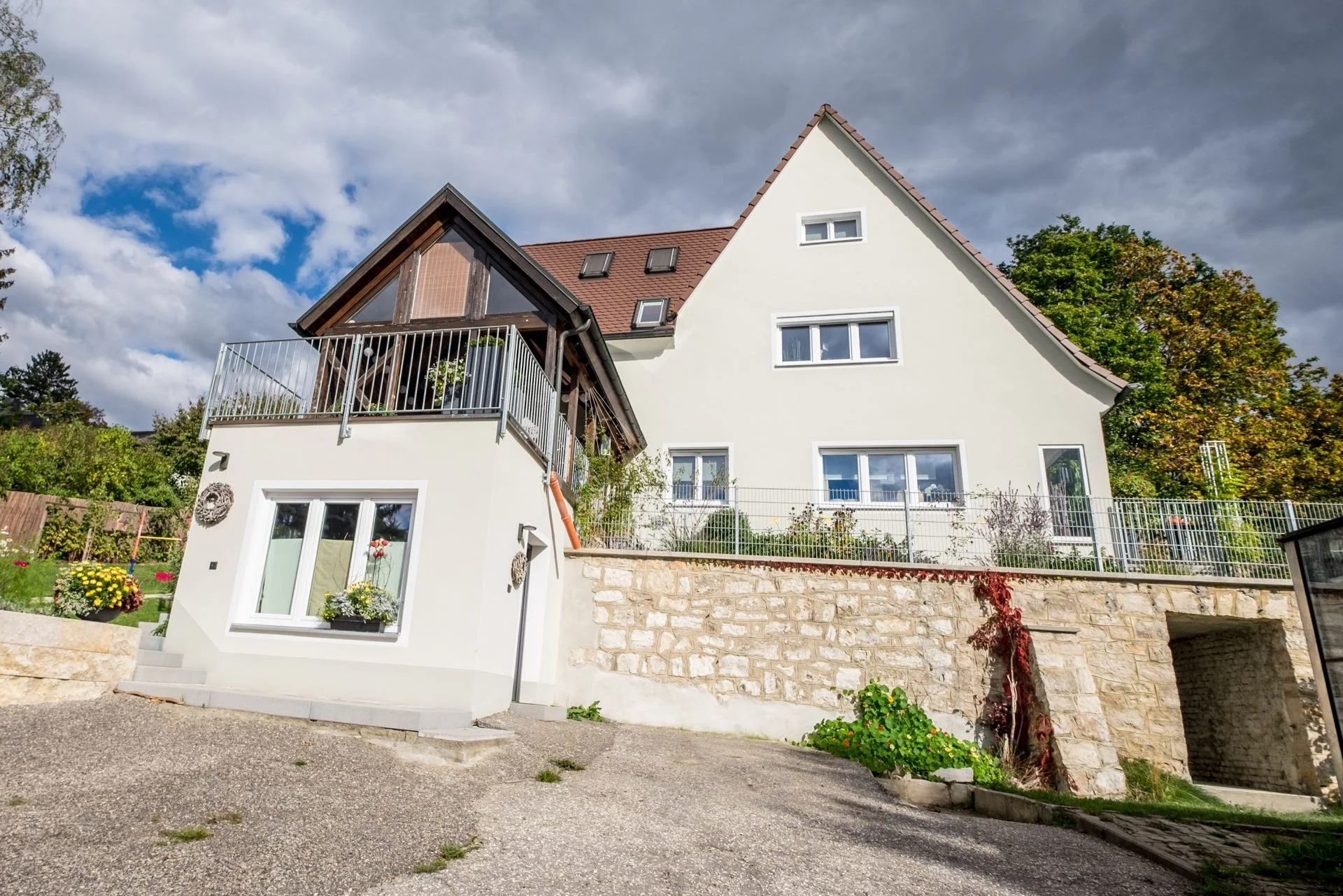 Ein modernes, mehrstöckiges Haus mit weißen Wänden, grauen Fenstern und einem roten Satteldach. Das Haus hat einen Balkon mit Geländer und ist auf einem erhöhten Grundstück gebaut, das mit einer Steinmauer und Zäunen abgegrenzt ist. Der Garten ist be