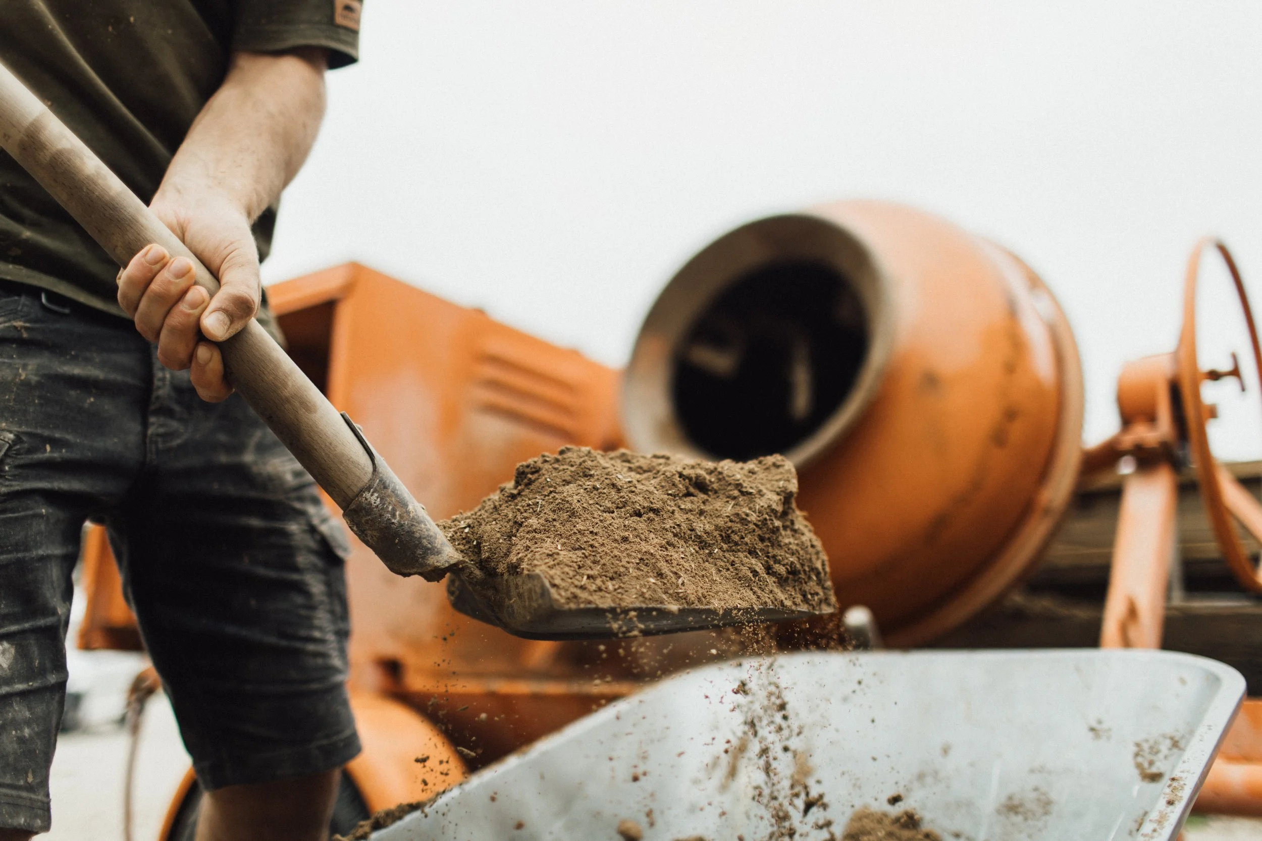 Person beim Betonmischen mit Schaufel, Betonmischer im Hintergrund.