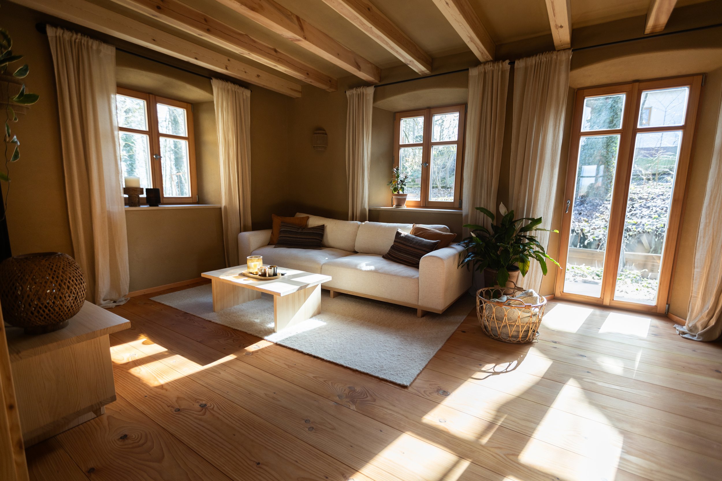 Gemütliches Wohnzimmer mit Holzboden, großen Fenstern und einem weißen Sofa, dekoriert mit Kissen, einem kleinen Couchtisch mit Kerzen, Pflanzen und Gardinen.