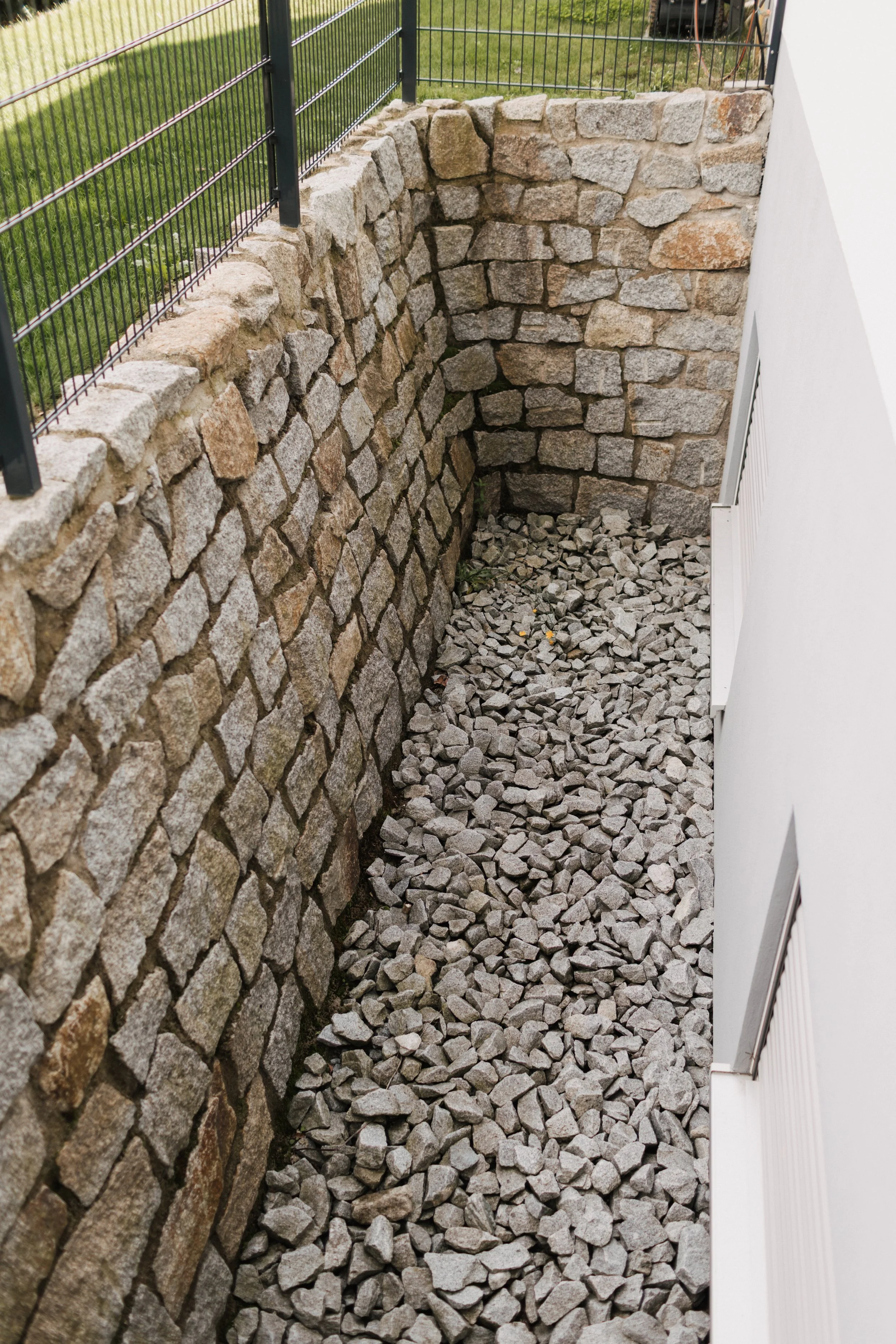 Ein abgeschlossener Innenhof mit einer Steinmauer, einer weißen Wand mit Fenstern und einer Bodenfläche aus Kieselsteinen.