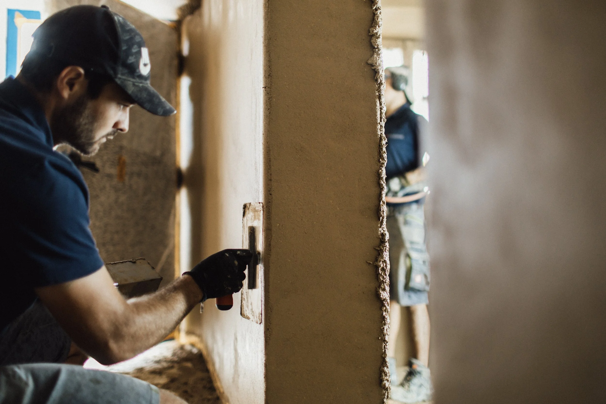 Ein Mann arbeitet an einer Wand, während im Hintergrund eine zweite Person steht. Der Mann mit Baseballcap nutzt ein Werkzeug, um die Wand zu bearbeiten.