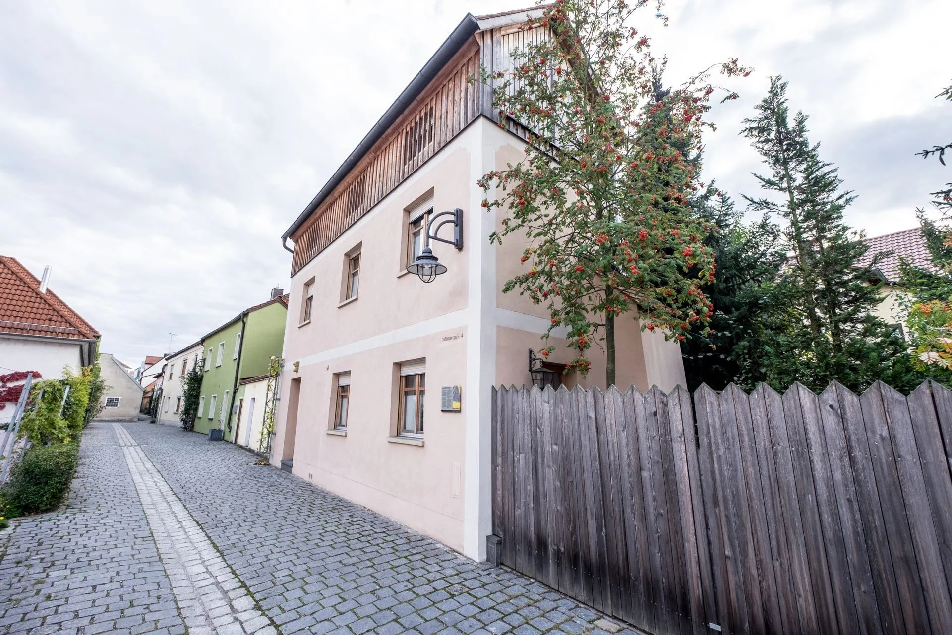 Ein mehrstöckiges Haus an einer schmalen Kopfsteinpflasterstraße, mit einer Holzzäune und einem Baum mit roten Beeren im Vordergrund.