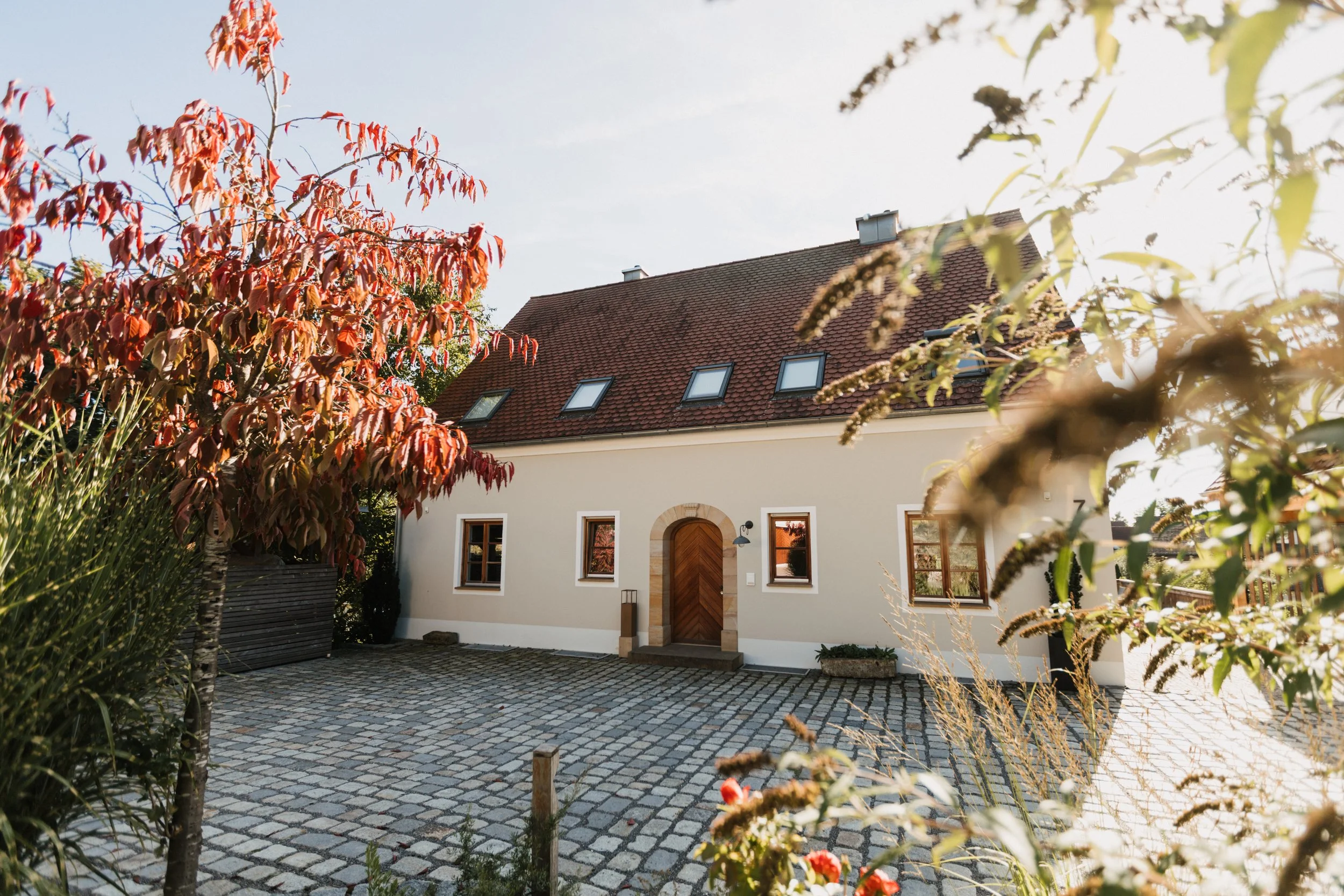 Ein Haus mit roten Dachziegeln, weißen Wänden und mehreren Dachfenstern, umgeben von Büschen und Bflumen im Sonnenschein.