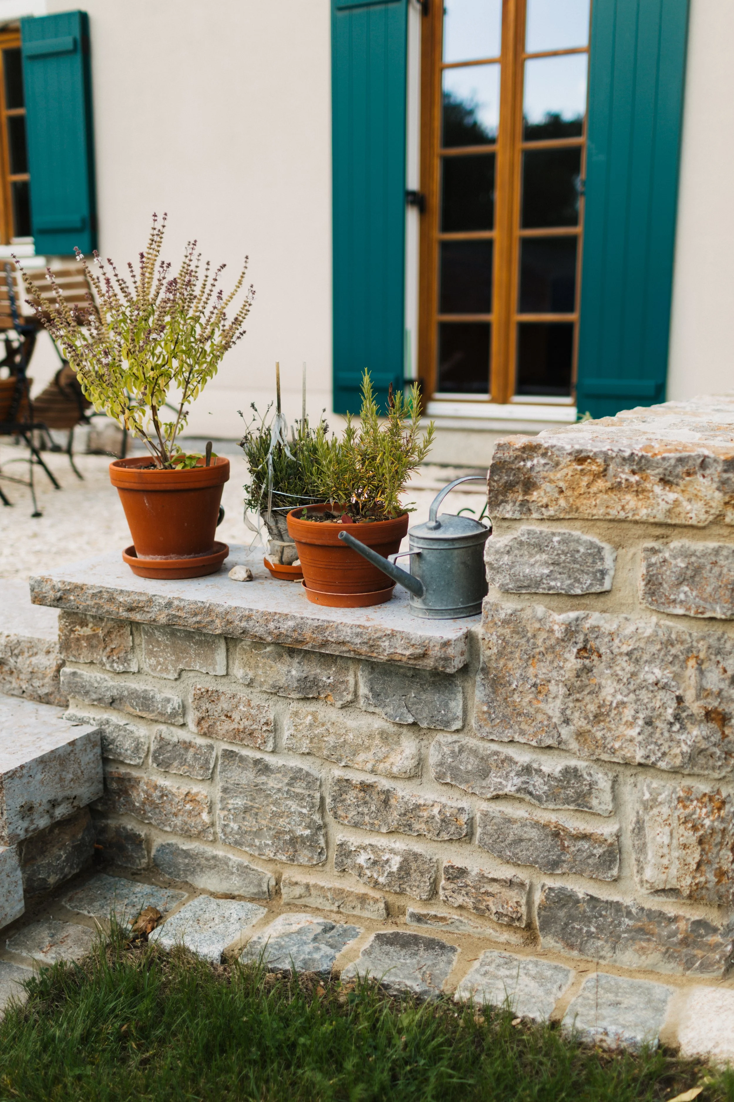 Blumentöpfe mit Kräutern auf einer Steinmauer vor einem Haus mit blauen Fensterläden.