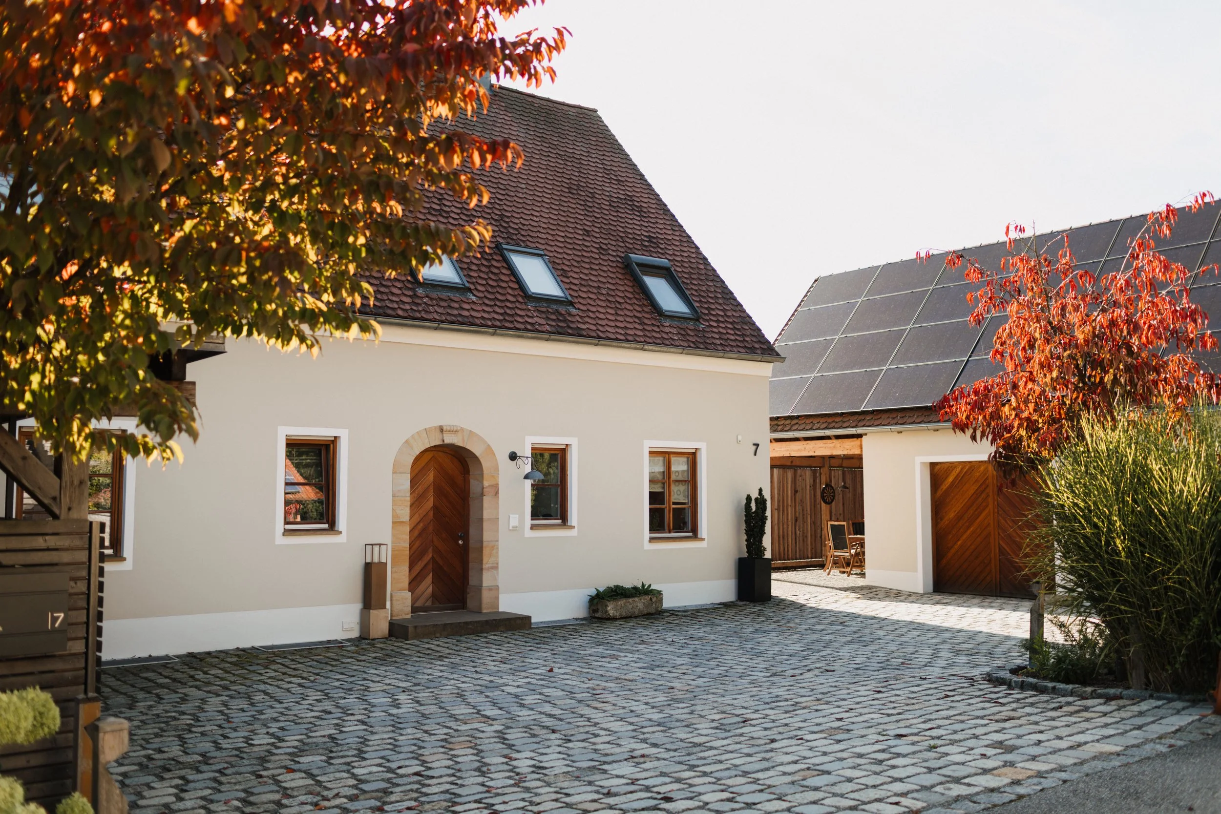 Häuser im ländlichen Stil mit einem gepflasterten Innenhof, eines mit weißer Fassade, rotem Ziegeldach und einem Holzportal, daneben ein Gebäude mit Solarmodulen auf dem Dach, umgeben von Herbstbäumen