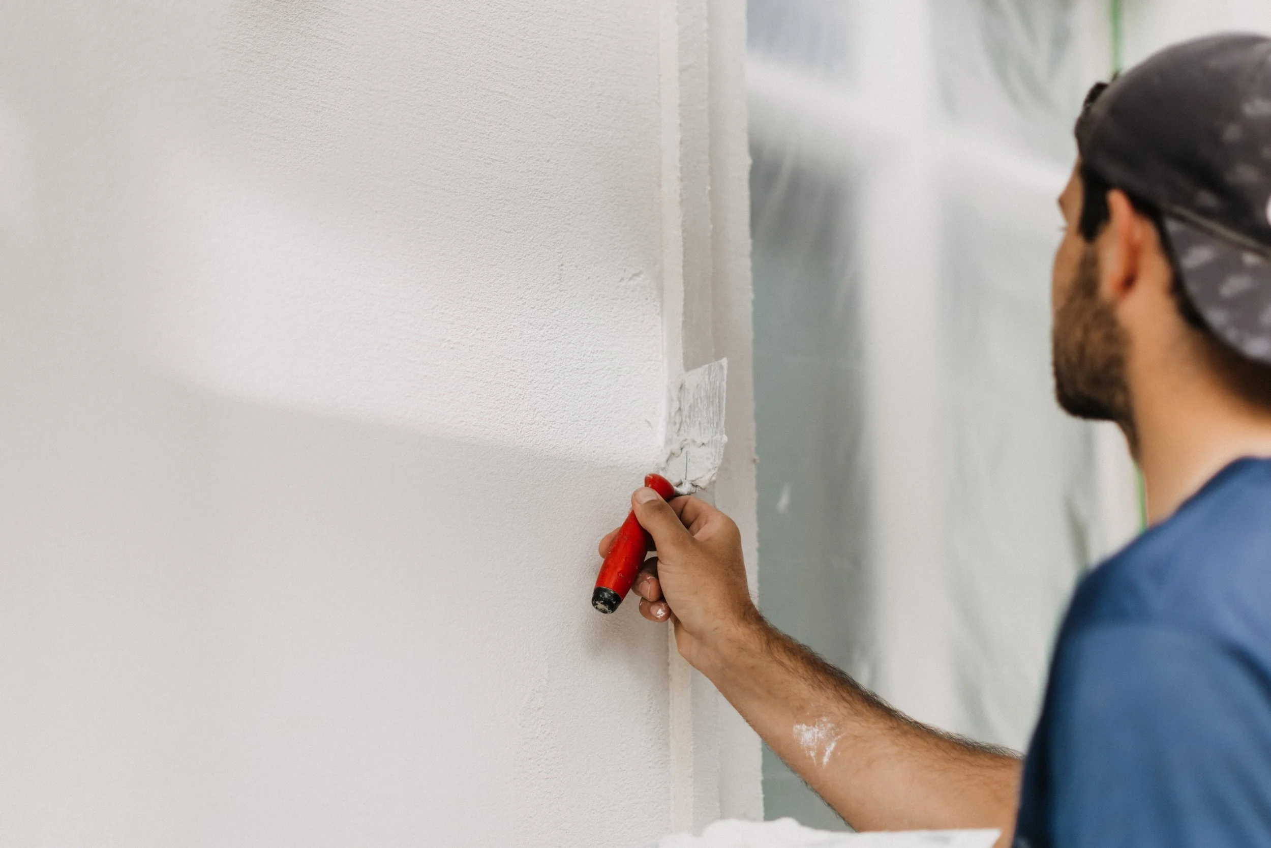 Ein Mann arbeitet mit einem Spachtel an einer weißen Wand, wahrscheinlich beim Tapezieren oder Glätten der Wand.