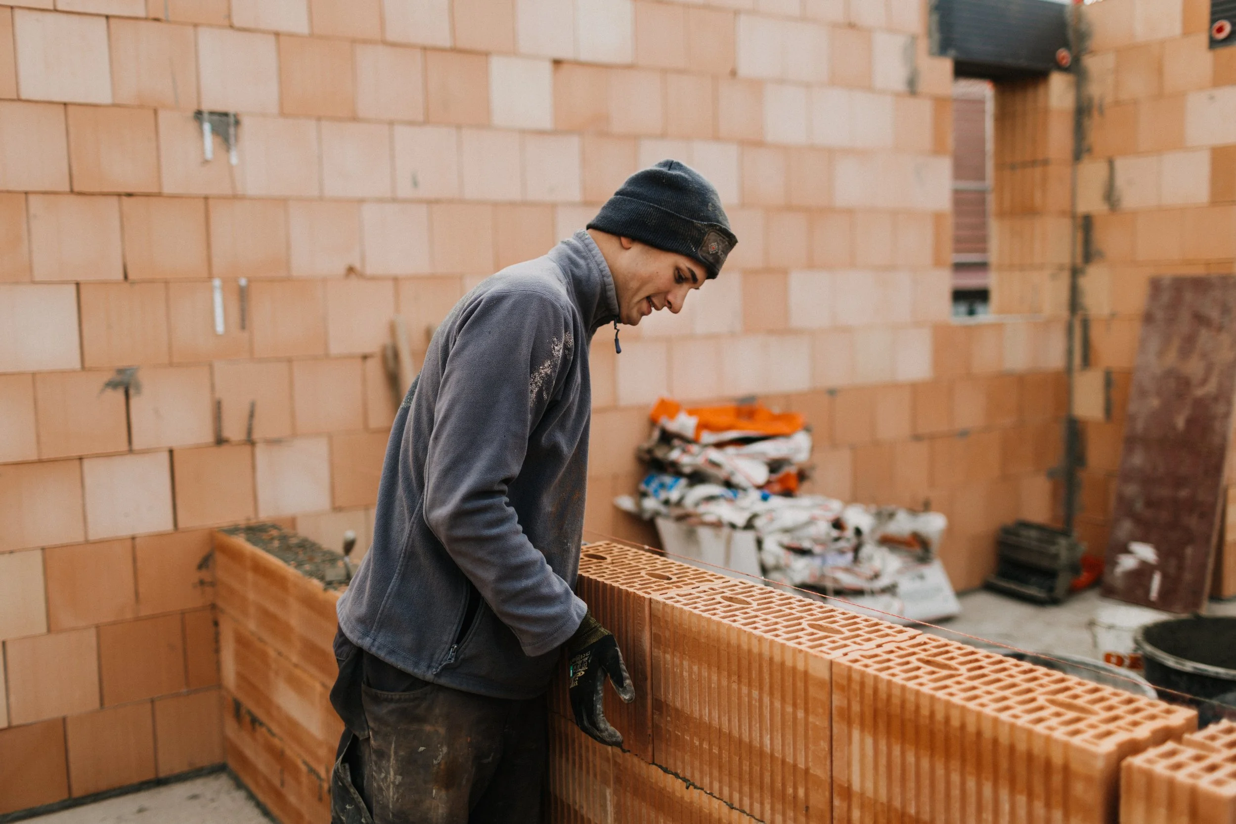 Mann beim Maurern beim Bau einer Ziegelmauer. Er trägt dunkle Arbeitskleidung und einen schwarzen Mützen. Im Hintergrund liegen Baumaterialien und Ziegel aufgestapelt.