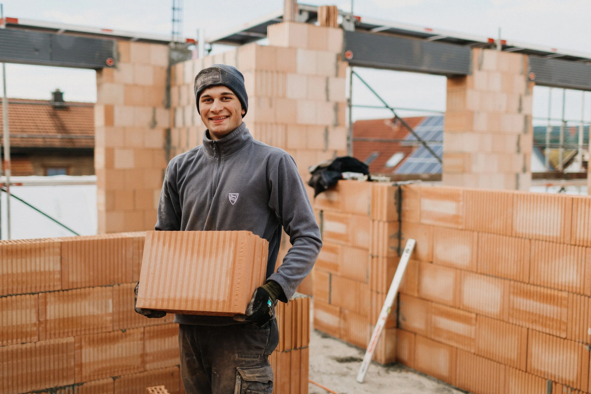 Ein junger Mann trägt einen Backstein und lächelt auf einer Baustelle, im Hintergrund sind Mauern und das Skelett eines Gebäudes zu sehen.