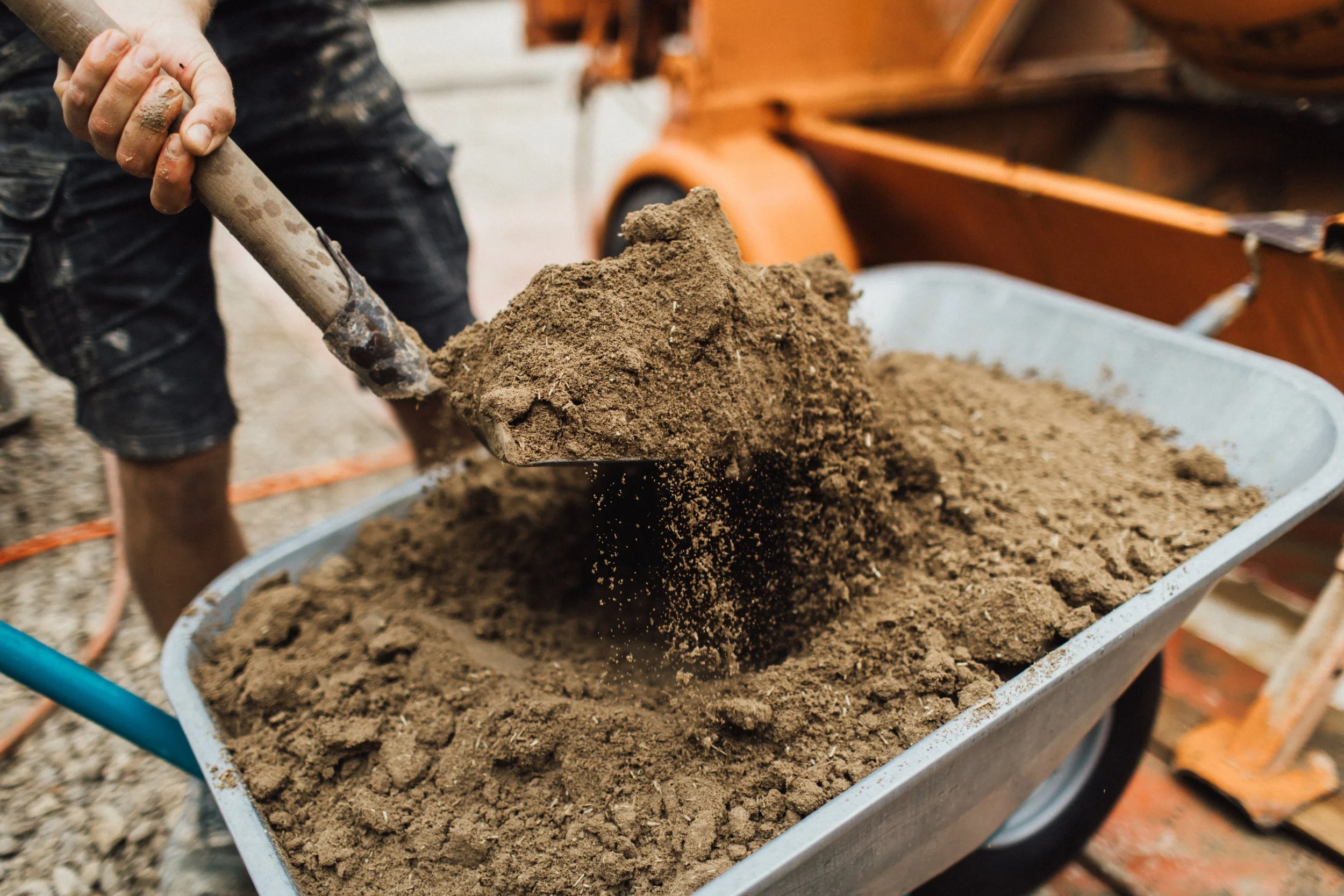 Person schaufelt Erde in eine Schubkarre, im Hintergrund ist ein alte landwirtschaftliche Maschine sichtbar.