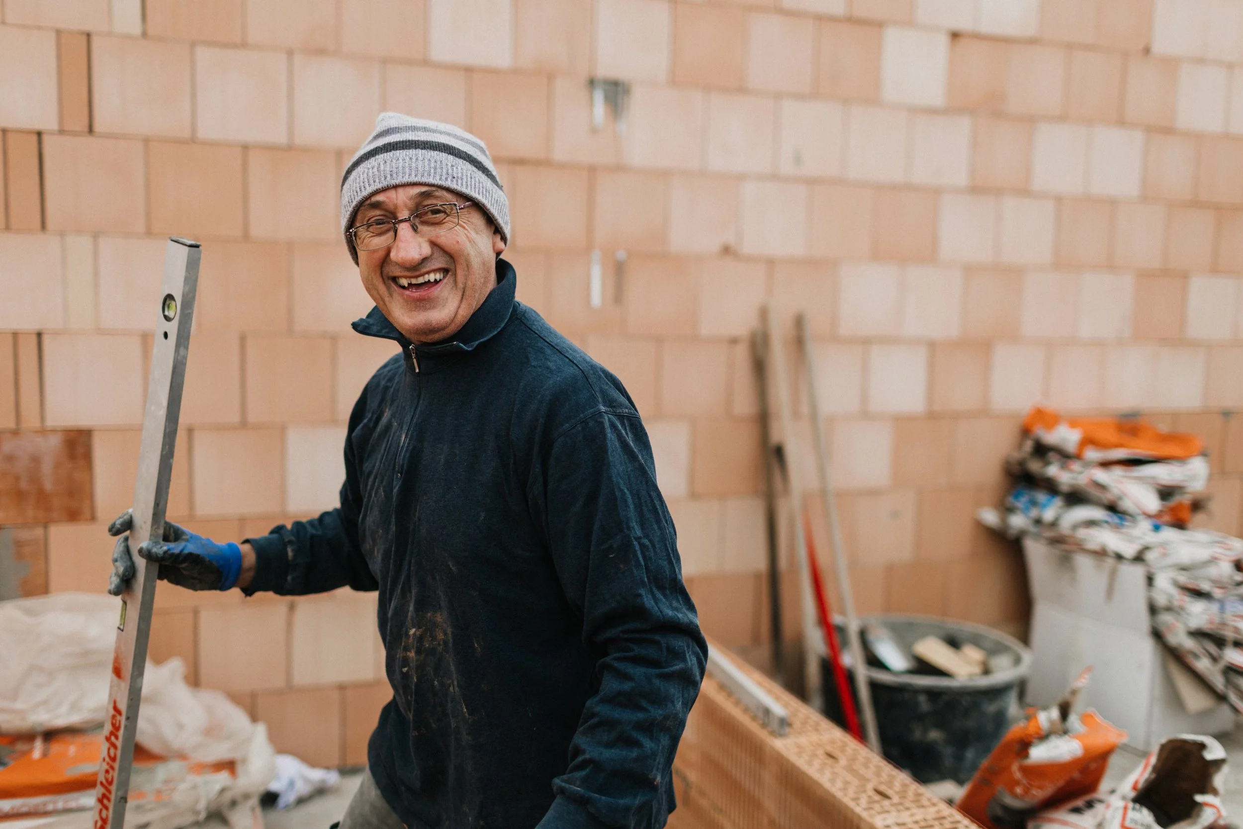 Ein lachender Mann mit Brille, grauem Mütze und dunklem Pullover beim Bauarbeiten, hält eine Wasserwaage in der Hand. Im Hintergrund sind Ziegelwände und Baumaterialien sichtbar.