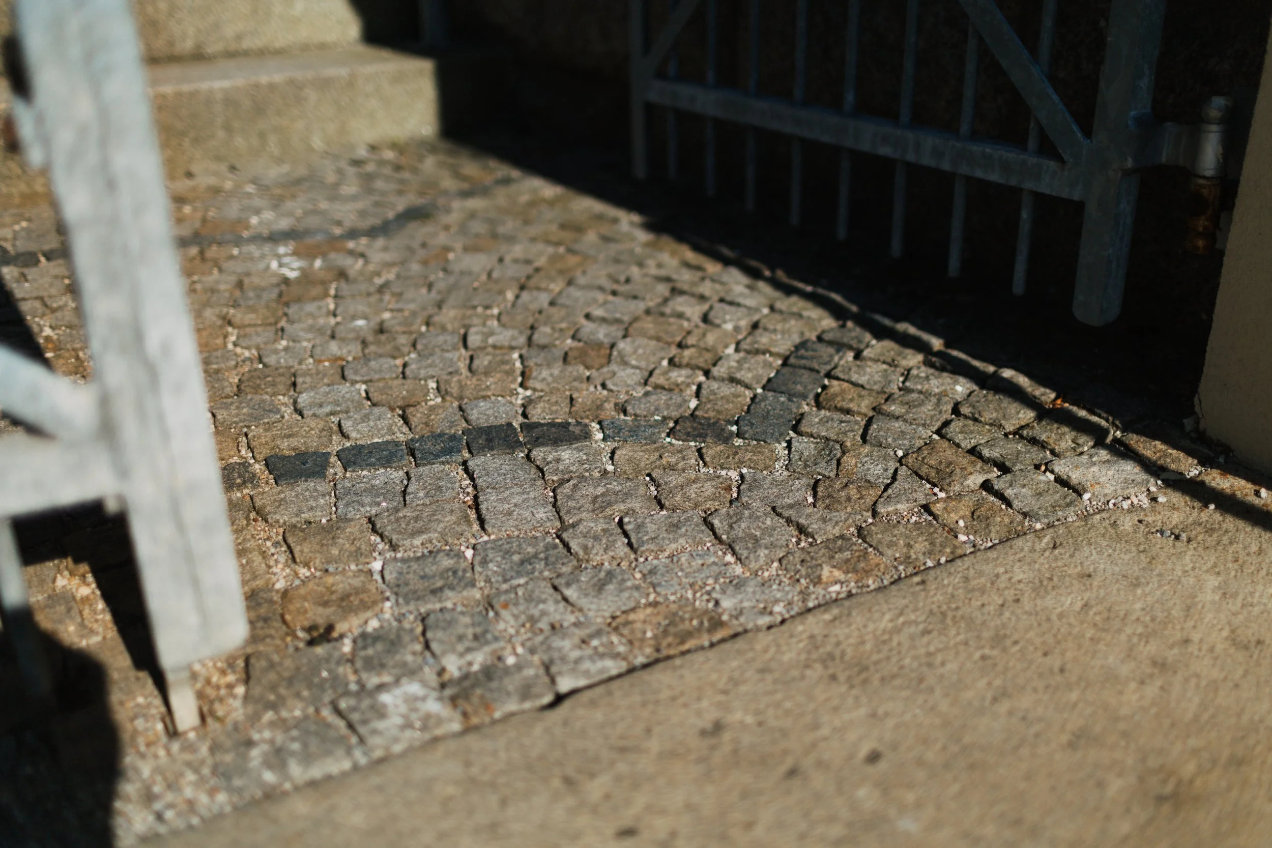 Boden aus kleinen, unregelmäßigen Steinquadern mit Schattenwurf, am Eingang einer Tür oder eines Zauns.