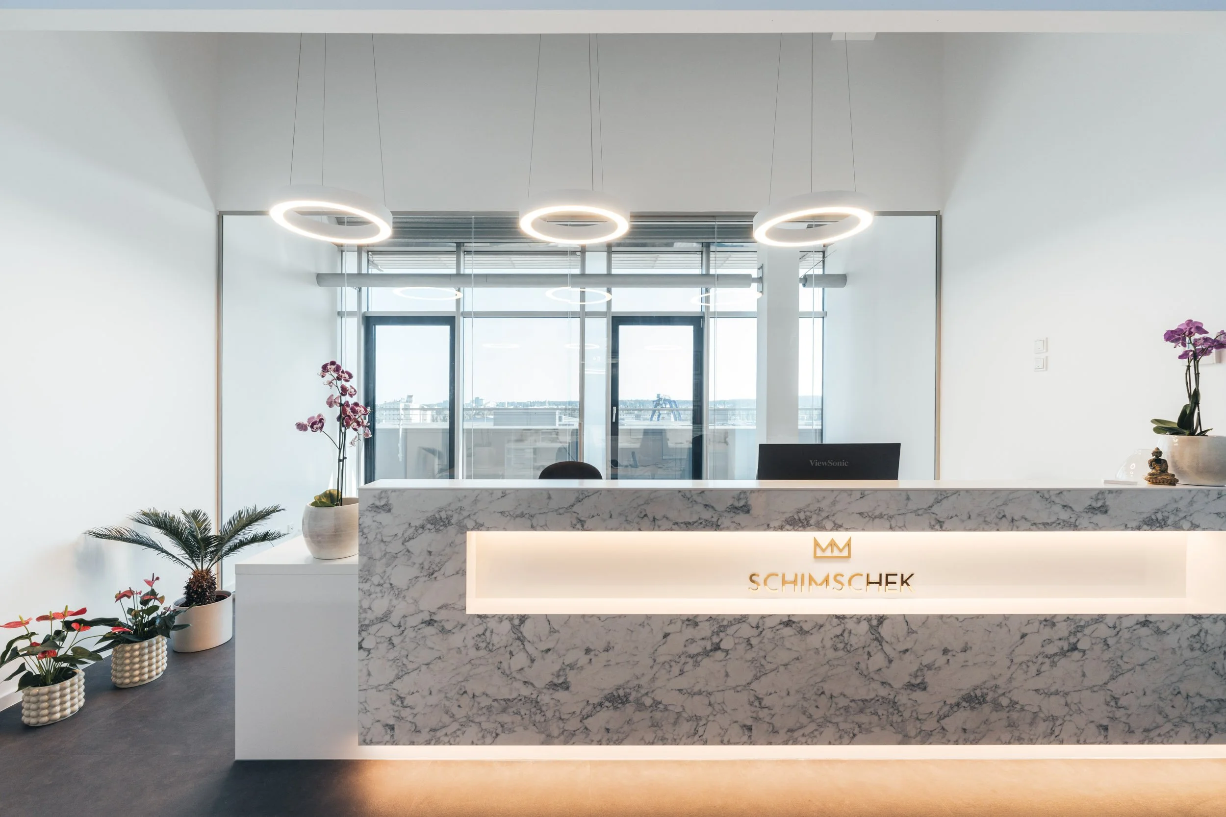 Modern reception desk with marble finish and backlit logo, surrounded by potted plants, in an office lobby with large windows and a city view.