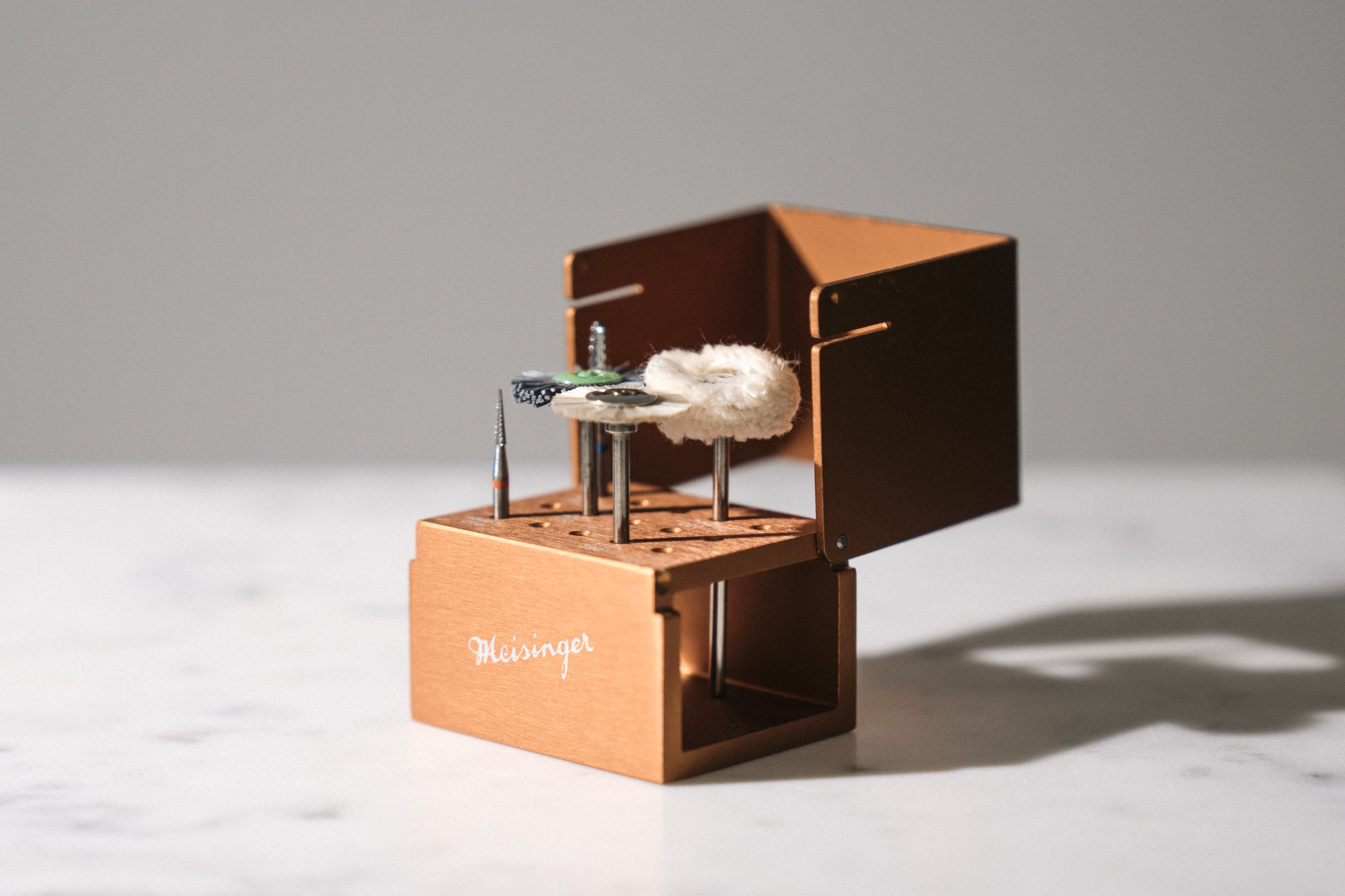 Close-up of a small open wooden box with sewing supplies, including a sewing pin, a spool of green thread, a fluffy white piece, and metal objects, on a white surface with a plain gray background.