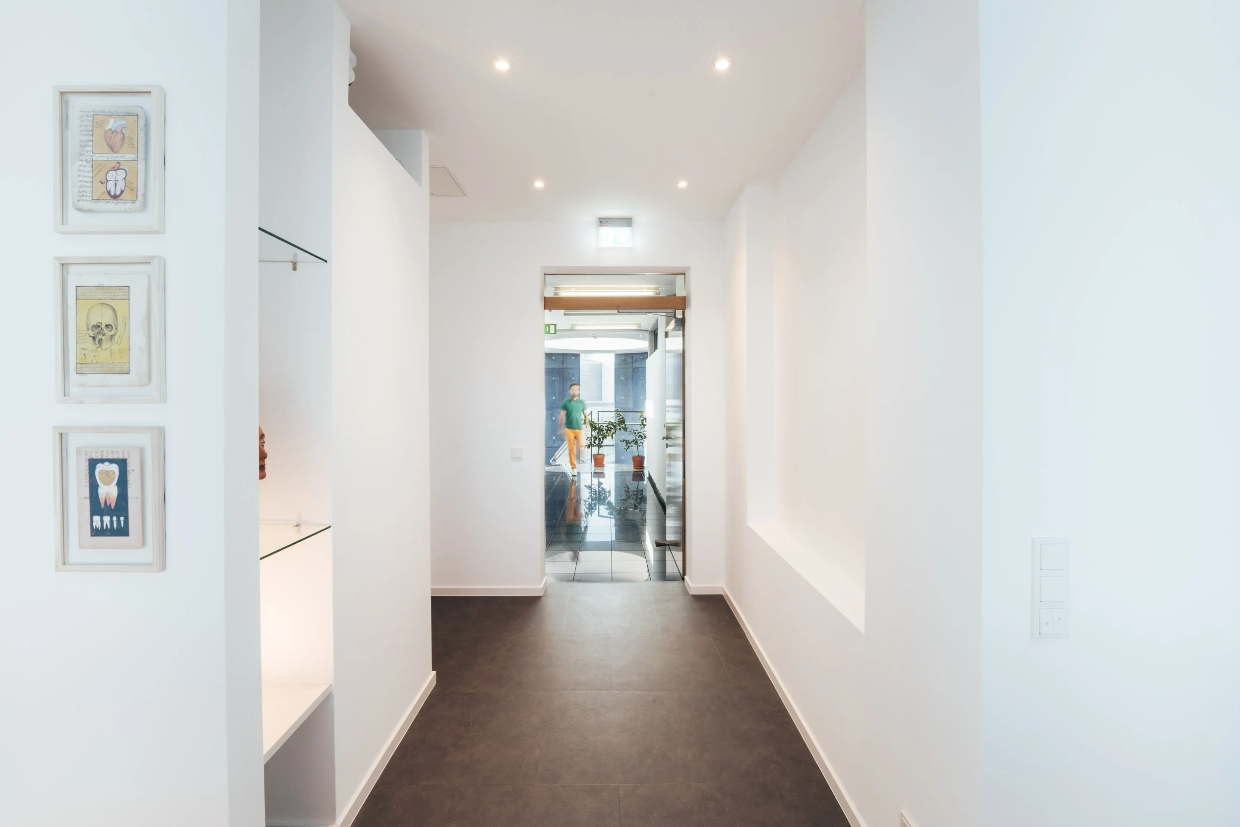 Clean, modern hallway in a building with white walls, dark flooring, and ceiling lights. Potted plants are visible through the glass door at the end.