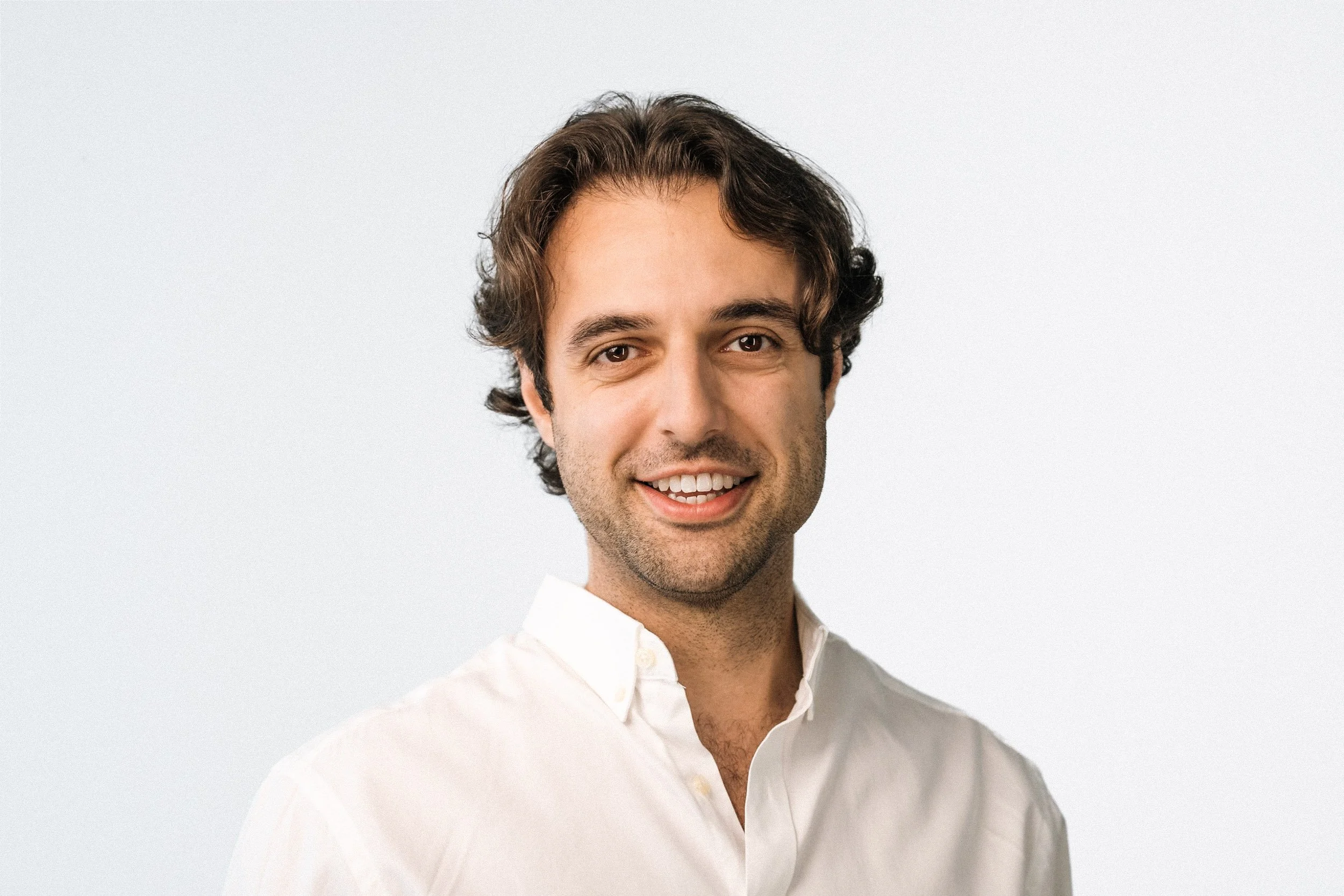 A smiling man with wavy brown hair wearing a white shirt against a plain light gray background.