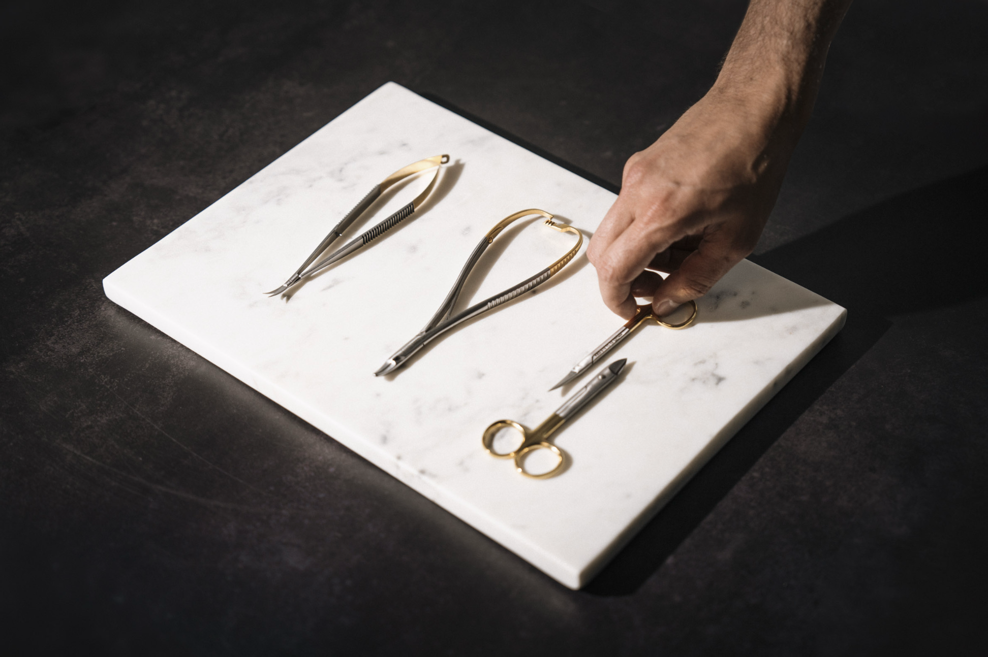A hand arranging gold and silver surgical scissors and forceps on a white marble cutting board.