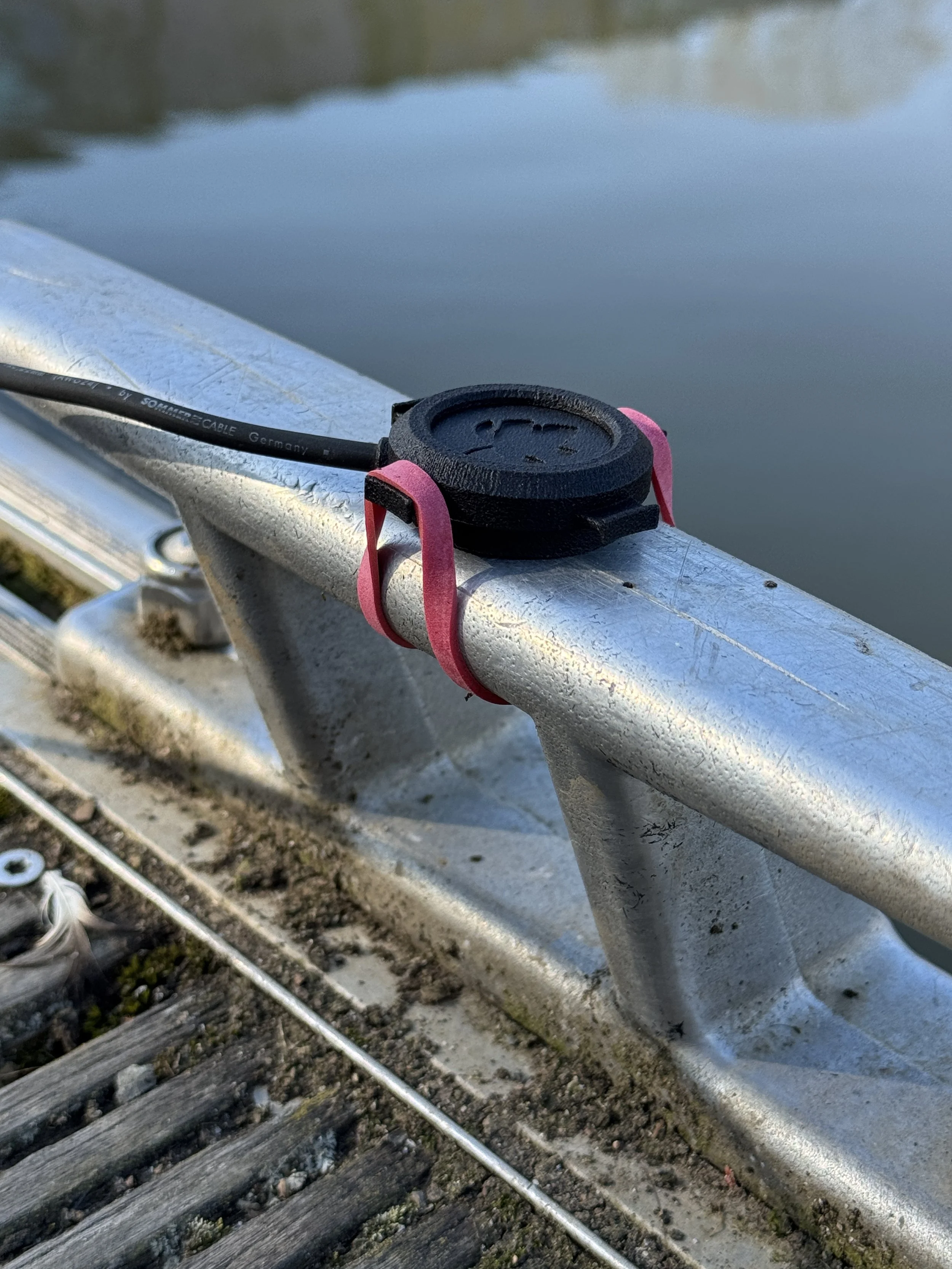 Close-up of a boat or dock railing with a black plastic cover, attached with red rubber bands, and a black cable running through it, near a body of water.