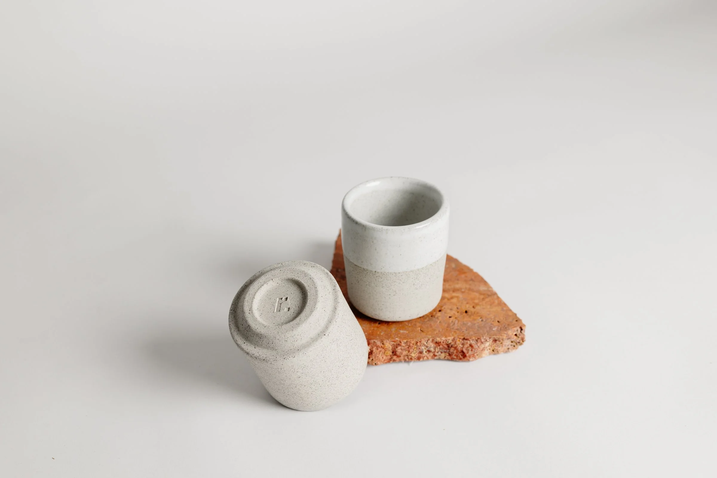 Two minimalist ceramic cups, one upright and one upside down, placed on a small piece of brick on a white surface.