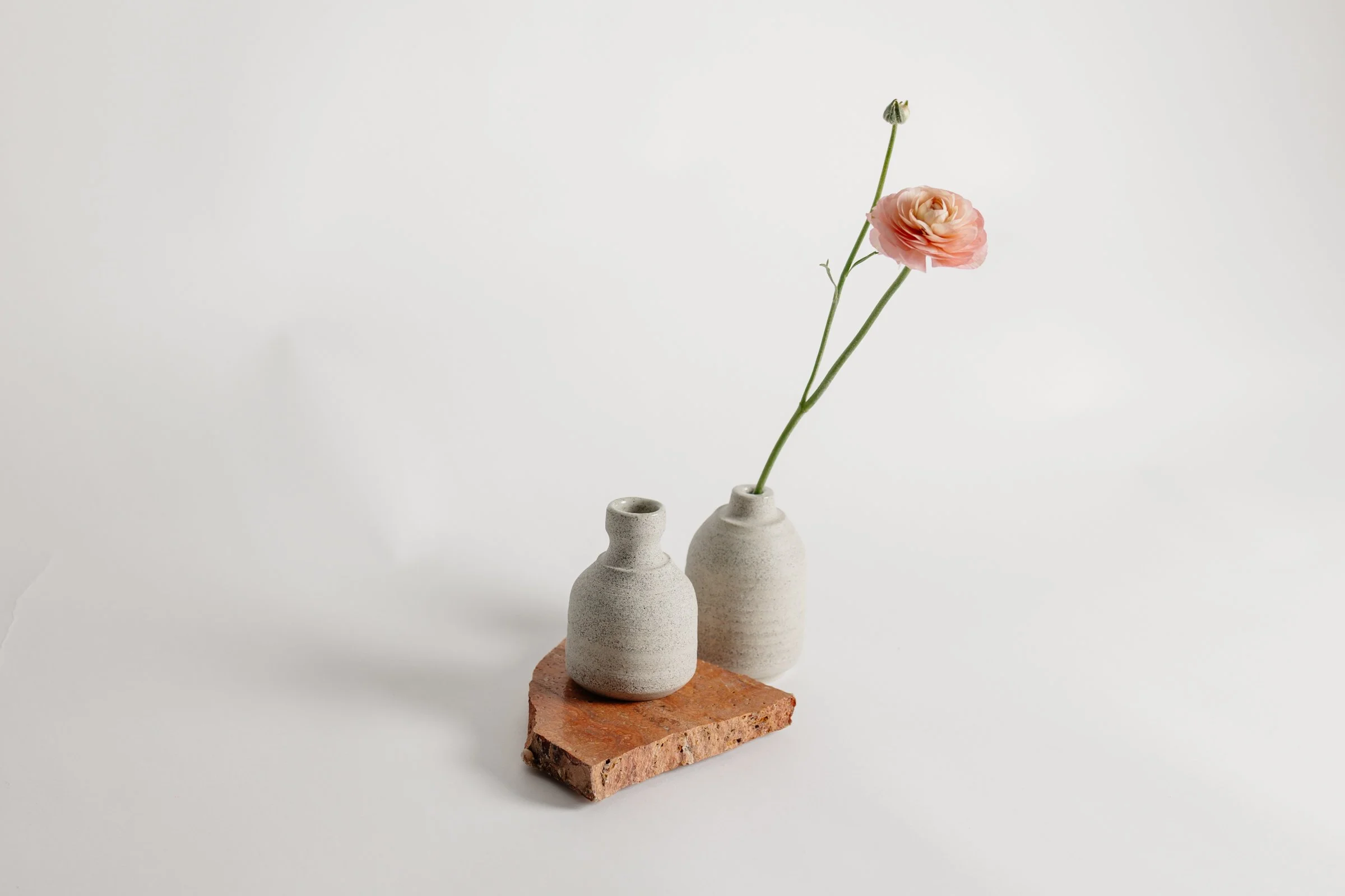 Two small ceramic vases, one with a pink flower, on a brick slab against a white background.
