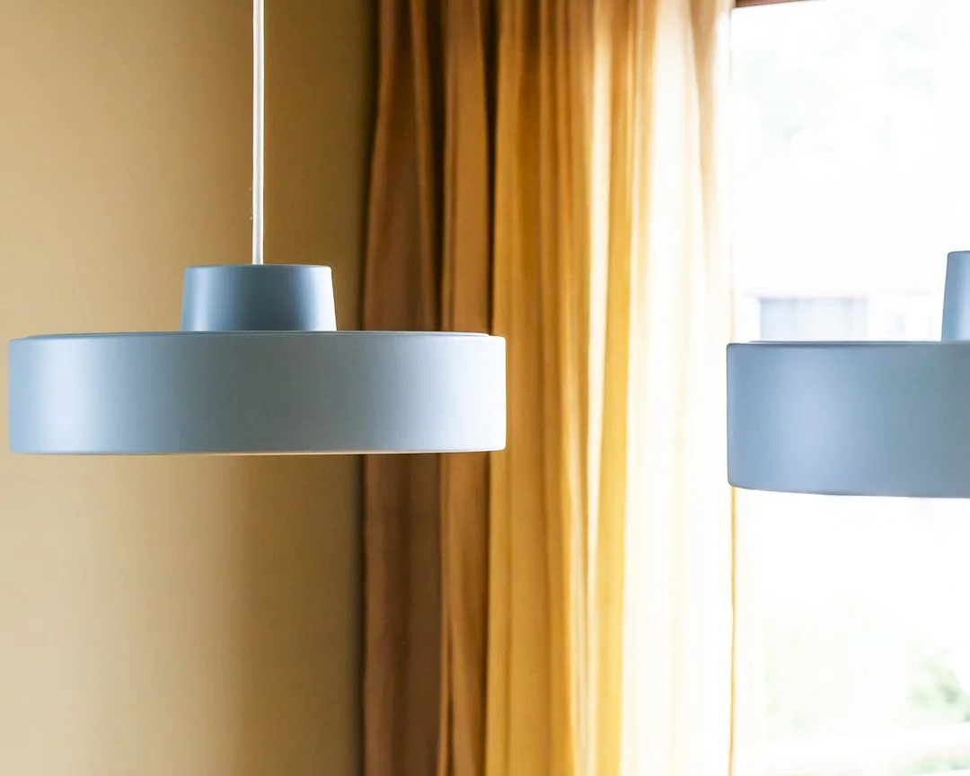 To moderne takpendler i lyseblåfarge henger fra taket foran store vinduer med gule gardiner.