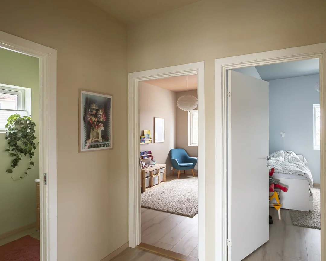Utsikt fra en gang med tre dører inn til tre forskjellige rom, ett med grønne vegger og en plante, ett med et blått sittehjørne, og ett med et barnerom med seng og leker.