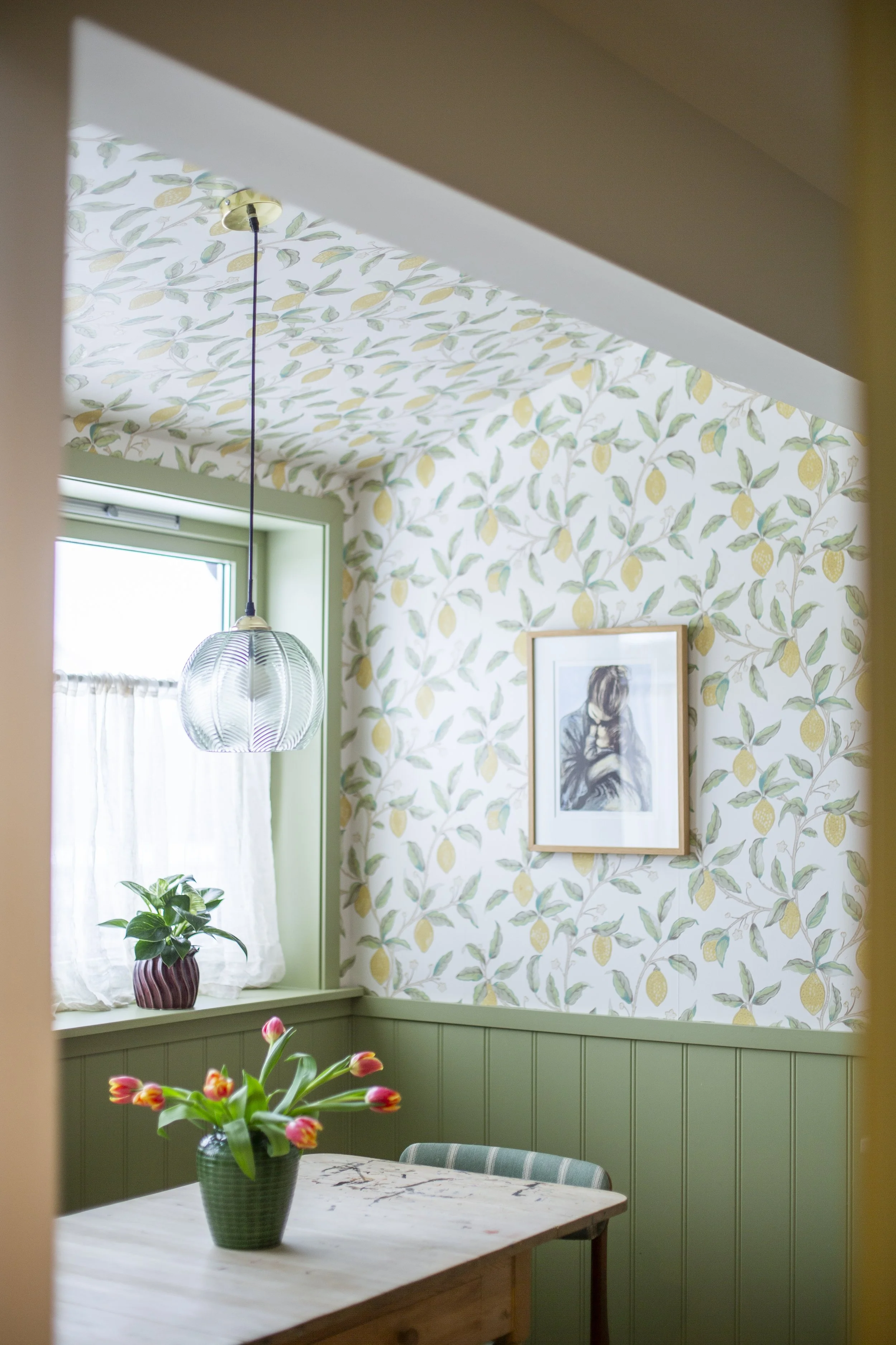 Et lyst kjøkken med grønne veggbelter, en blomsterpotte med tulipaner på et trebord, en vinduskarm med en grønn plante i en krukke, og et bilde på veggen.