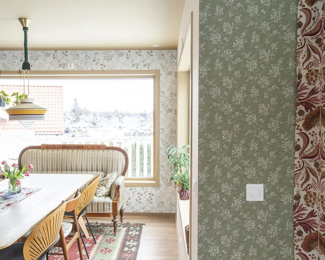 Et lys innendørsrom med et spisebord, stoler og en lenestol med puter, store vinduer med utsikt over nabolaget, dekorative tapeter på veggene og planter på en hylle. Kombinasjonen av mønstrede tapeter og treverk skaper en koselig atmosfære.