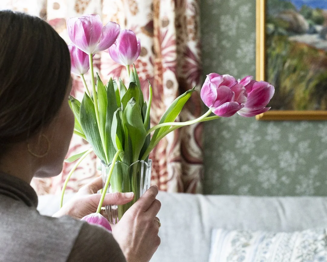 En person som holder en vase med rosa tulipaner innendørs, med gardiner og maleri i bakgrunnen.