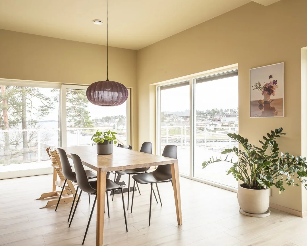Et lyst spiserom med et rektangulært trebord, seks svarte stoler, store vinduer med utsikt over et landskap, en grønne plante på bordet og en større plante i hjørnet, samt et bilde av blomster på veggen.