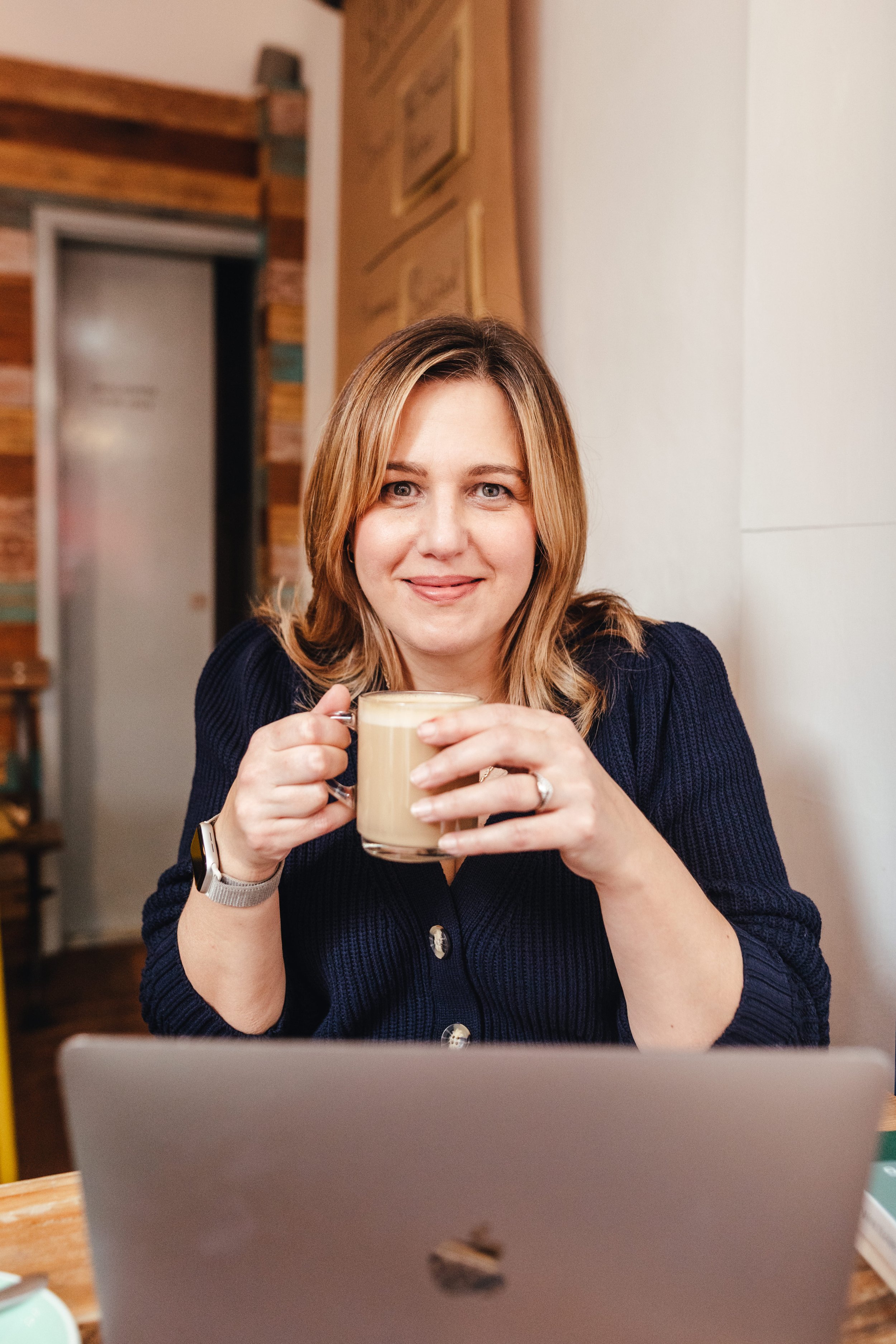 Michelle Jehu, founder of This Square Peg. A woman with shoulder-length blonde hair wearing a navy blue sweater, holding a coffee mug, sitting at a desk with a laptop, smiling at the camera in a cosy, wooden interior space.