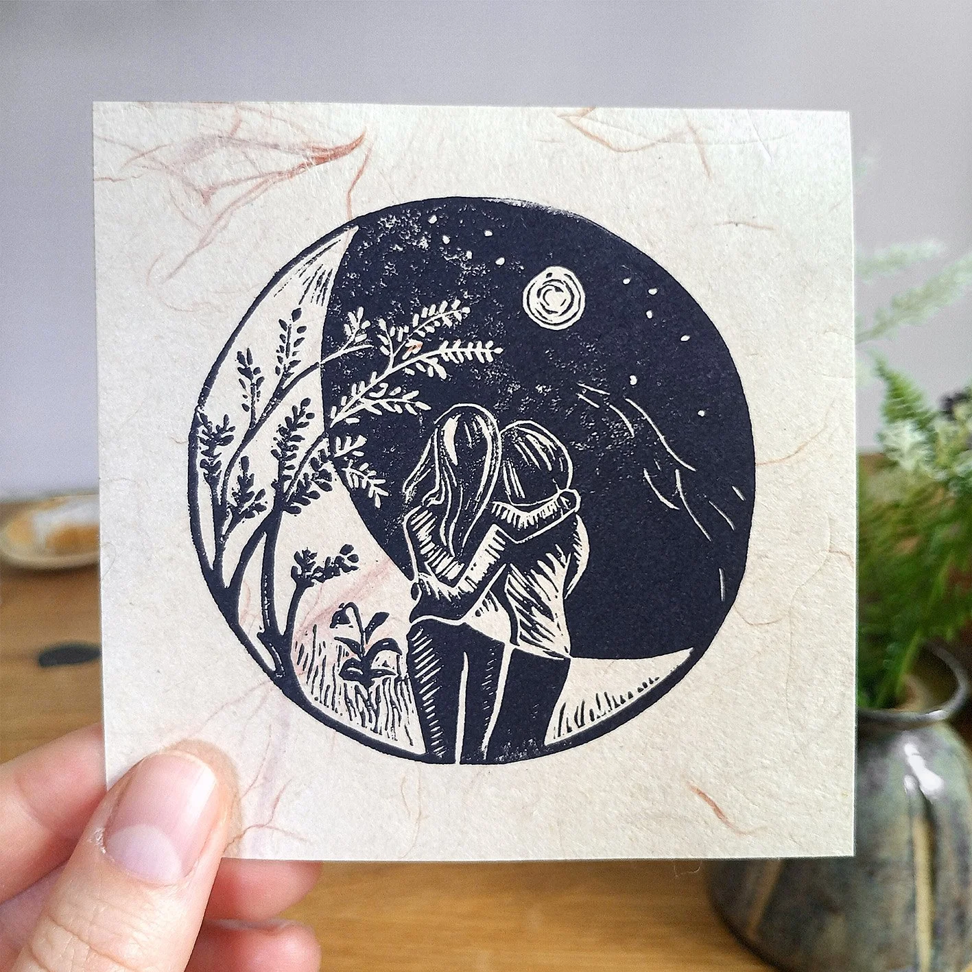 eine Hand die einen Mini Linoldruck hält, auf dem eine Nachtlandschaft mit zwei Frauen zusehen ist, die im Mond stehen und sich in den Armen halten.