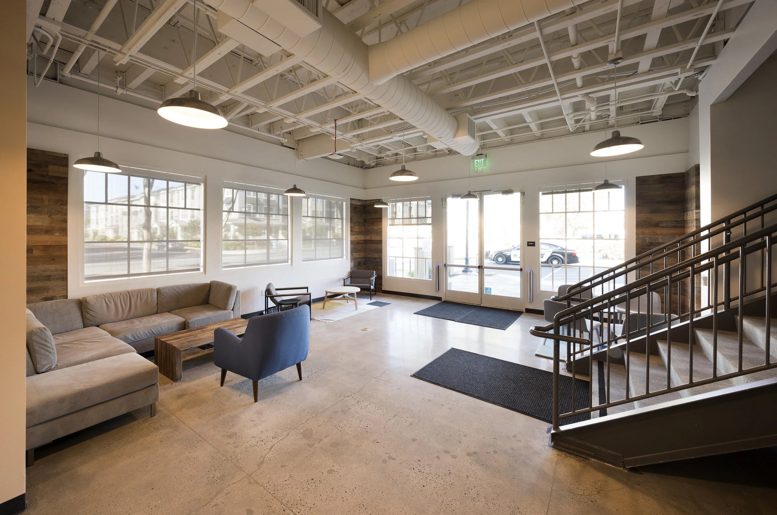 A spacious, modern lobby area with large windows, a beige sectional sofa, and a mix of chairs arranged near the windows. The ceiling has exposed ductwork and pendant lighting. The entrance features glass doors with an exit sign above.