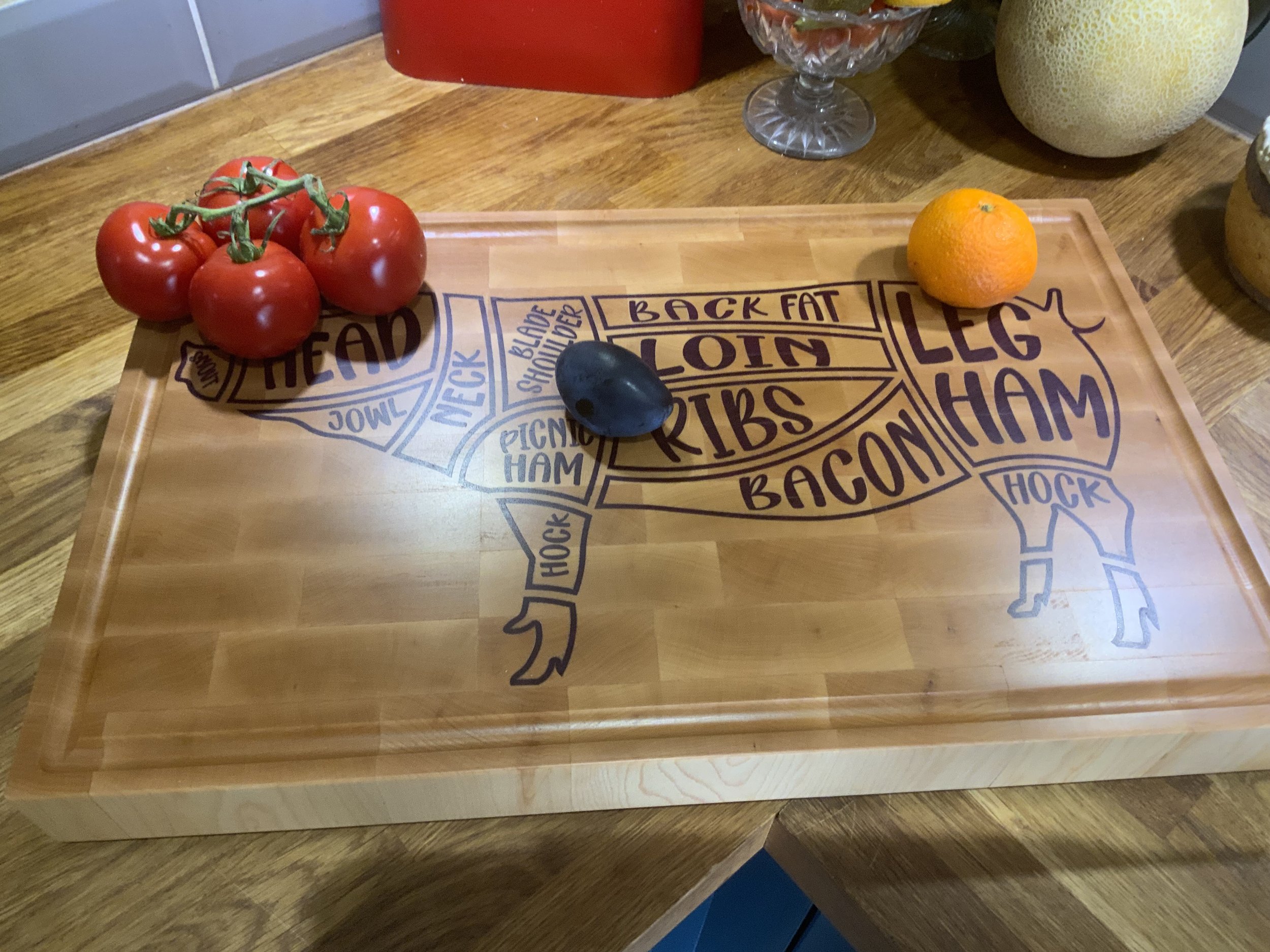 A wooden butcher block with a design of meat cuts, sitting on a wooden table, with a bunch of tomatoes on the left, a black stone in the middle, and an orange on the right.